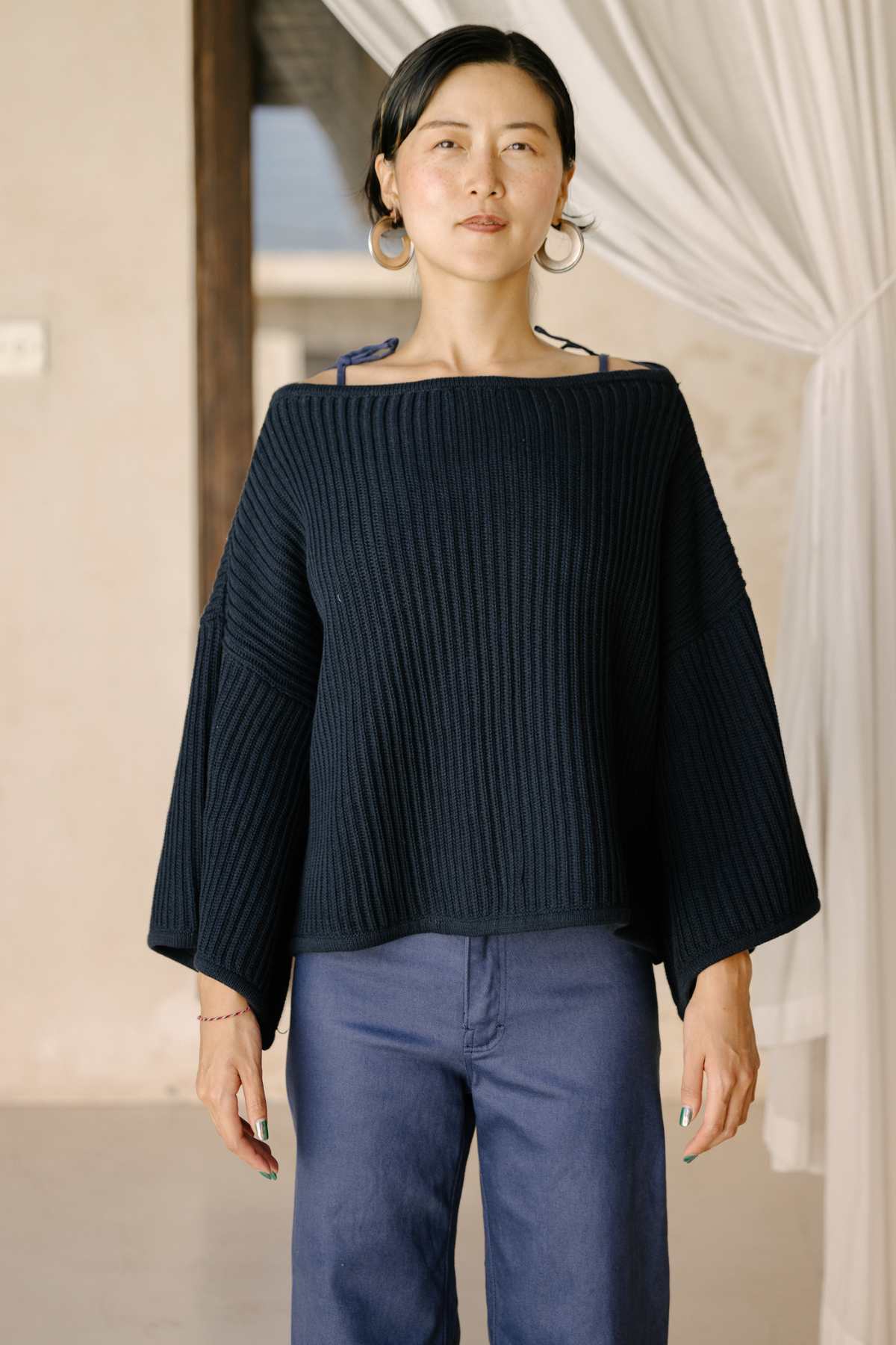 A person with tied-back dark hair stands indoors, wearing the Spirit Top Sweater Knit by Myrah Penaloza, a luxurious black ribbed cotton sweater featuring wide sleeves. They have paired it with blue pants and are positioned in front of a light-colored wall, with sheer curtains partially visible in the background.