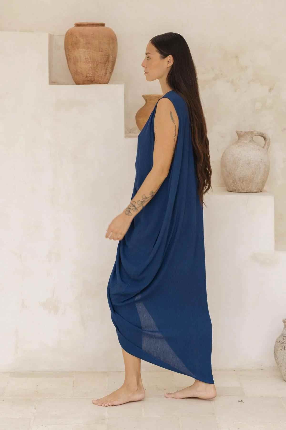 A woman with long dark hair and visible tattoos on her arm is walking barefoot on a light-colored surface. She is wearing a flowing blue Virgo Luna Dress by Myrah Penaloza, featuring a high neckline and sleeveless style. In the background, several ceramic vases are arranged on stepped surfaces.