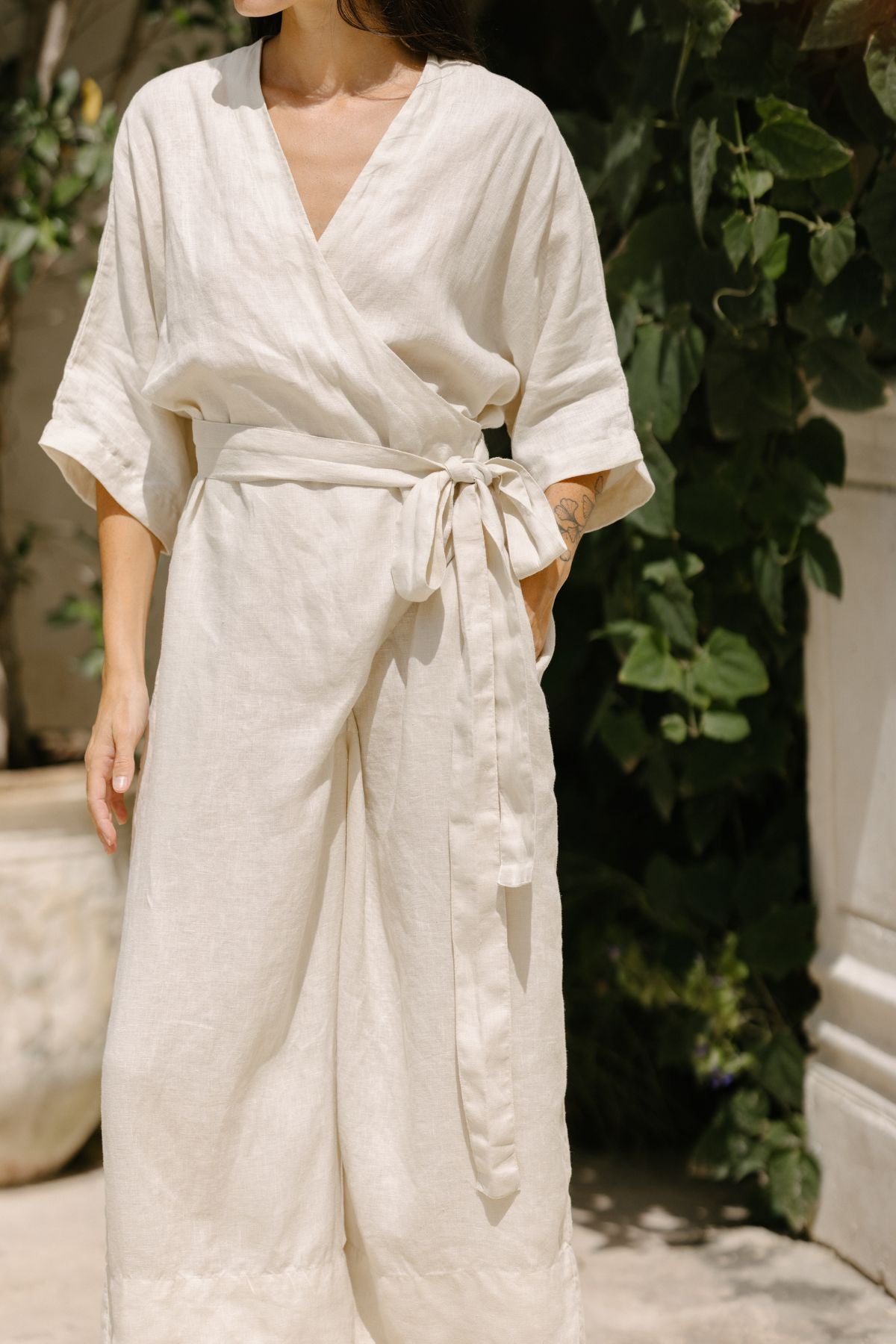 A person is wearing the Rey Linen Playsuit by Myrah Penaloza, a light-colored, loose-fitting outfit with a tied belt around the waist. The individual is standing outdoors with green foliage and a stone structure visible in the background, showing only their torso and part of their legs.