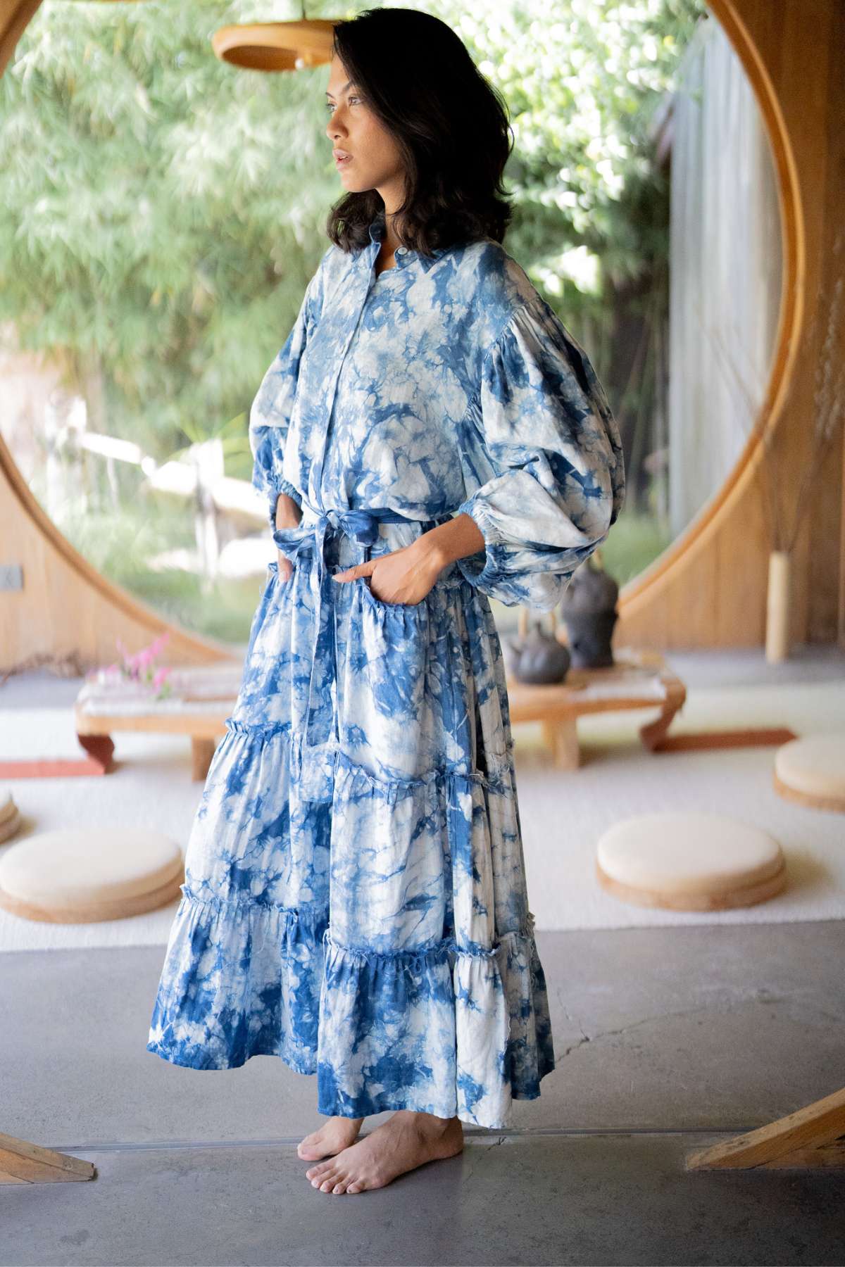 A woman stands barefoot in a room with a large circular window, wearing the Myrah Penaloza La Dama Long Sleeves Ruffles dress. This renaissance-inspired gown, hand tie-dyed in indigo hues and complete with a belt, beautifully complements the lush greenery outside and the round cushions scattered on the floor.