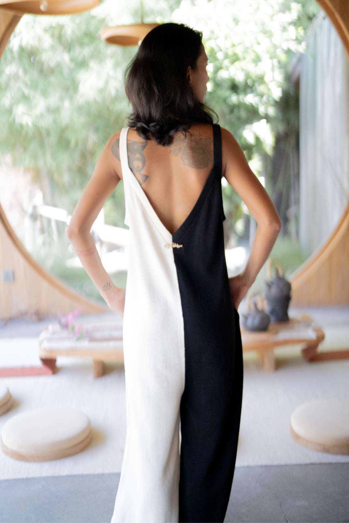 A person with shoulder-length hair stands with their back to the camera, adorned in a black and white Yin Yang Kundalini Knitted Playsuit by Myrah Penaloza. Visible tattoos decorate their shoulders, framed by a setting that includes a round window and minimalist indoor decor.