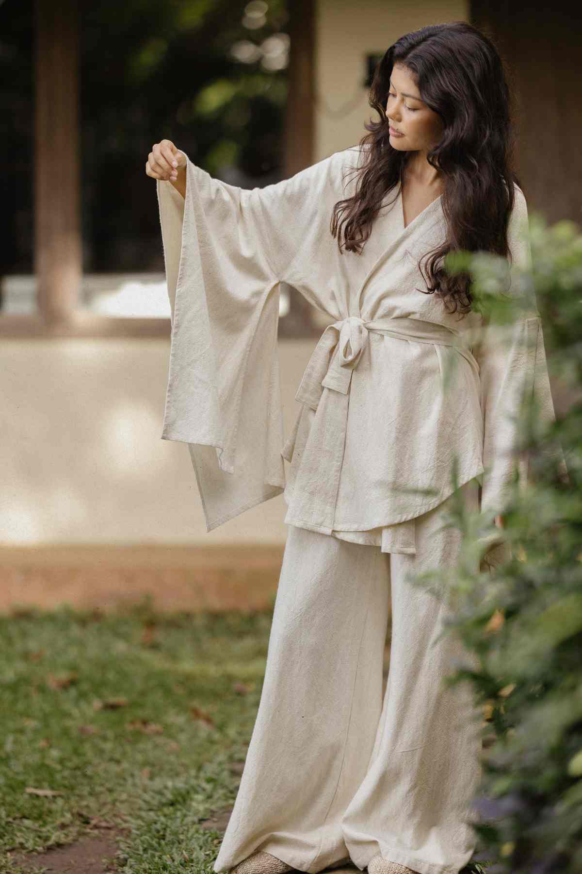 A woman with long, wavy hair stands outdoors wearing the Dewi Set (3 of a kind) by Myrah Penaloza, a flowing, cream-colored kimono-style outfit with bell sleeves and a tie at the waist. She holds one of the sleeves and gazes at it intently. This limited-edition piece stands out against the lush greenery and wooden windows in the background.