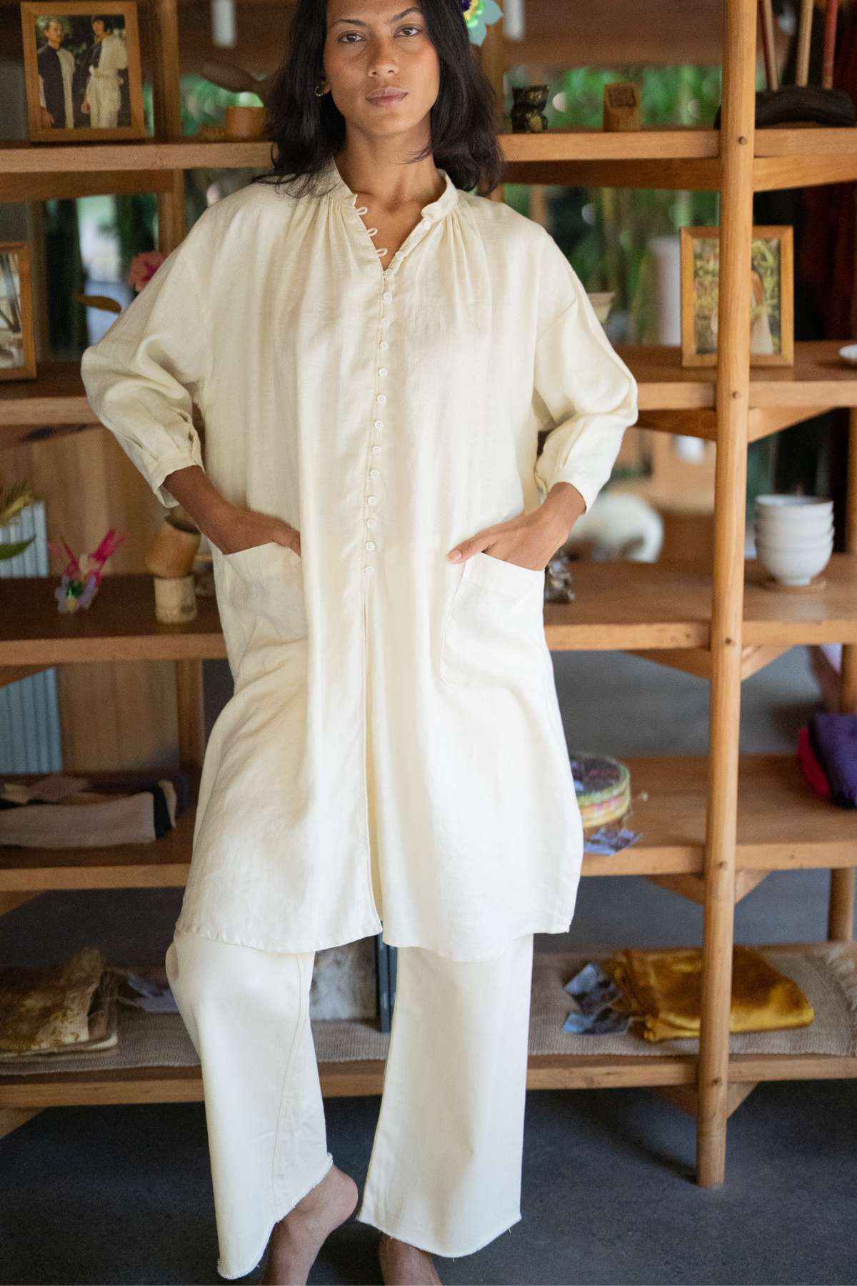 Dressed in a cream-colored Zazen Linen Top paired with matching pants, a person effortlessly exudes elegance indoors, reminiscent of the Frida Gown Original. They stand before a wooden shelf adorned with ceramic dishes and rolled textiles, creating a cozy and casual atmosphere.