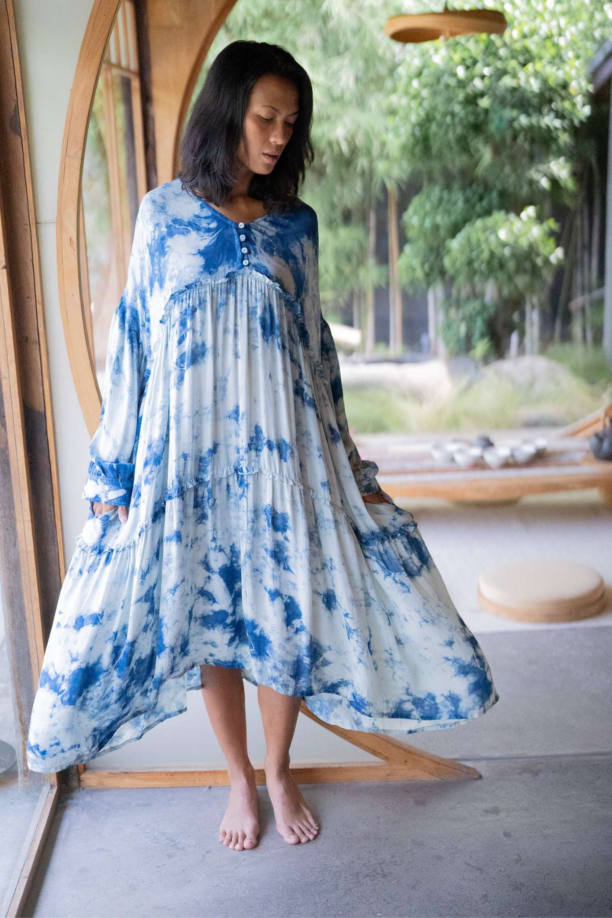 A woman stands indoors wearing an Indigo Eva Gown, crafted from 100% silk by Myrah Penaloza, featuring a blue and white tie-dye pattern with an oversized fit. She is barefoot, holding the sides of her dress. Behind her is a wooden and glass door with greenery visible outside, highlighting nature's touch.