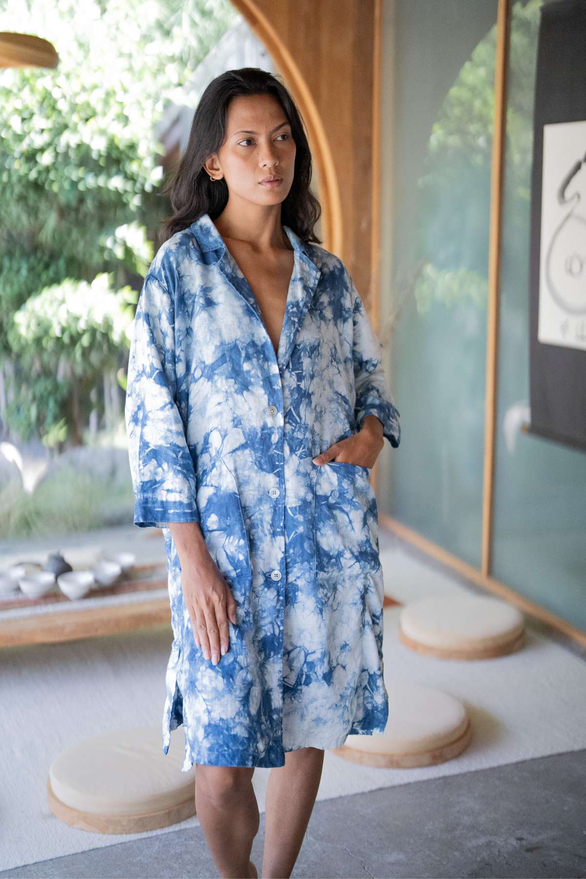 A person wearing a Myrah Penaloza Rubi Linen Jacket Indigo - Black Friday edition stands indoors, surrounded by circular cushions on the floor and a large window showcasing greenery in the background.