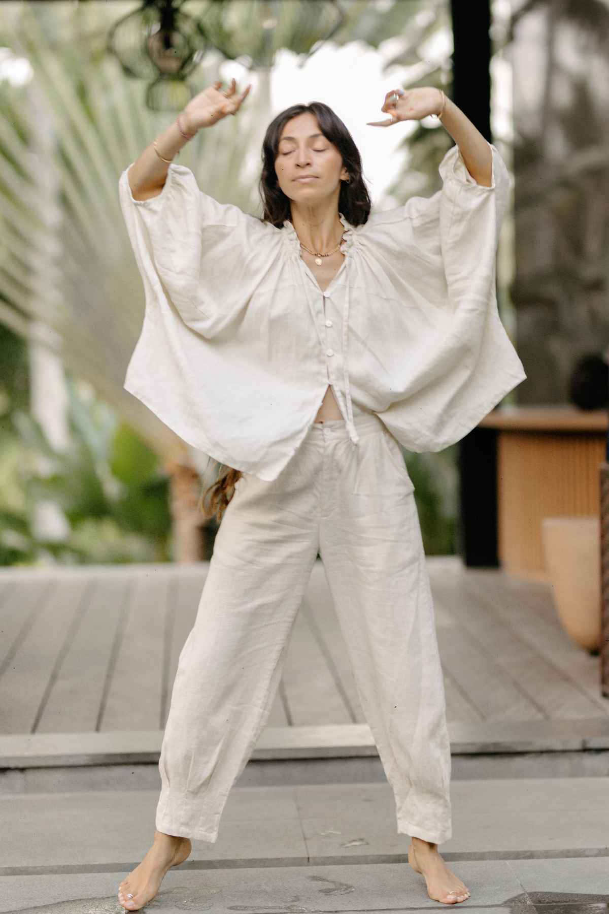 A woman wearing loose, flowy Myrah Penaloza Linen Chajin Pants and a white top is standing barefoot on a wooden deck. She has her eyes closed and her arms raised above her head. The background is blurred with green foliage and parts of a wooden structure visible, reflecting the serenity of her versatile wardrobe choice.