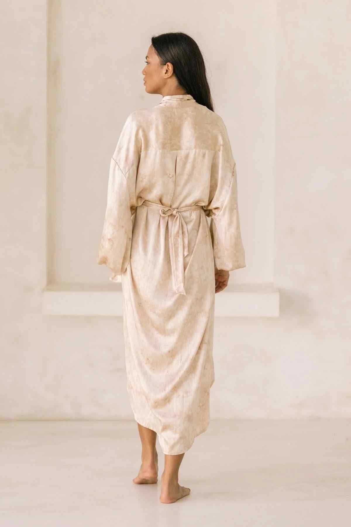 A person with long dark hair is standing barefoot, facing away from the camera. They are wearing a Silk Inanna Wrap Gown by Myrah Penaloza, a loose-fitting, light-colored robe tied at the waist with a sash. The background is neutral, emphasizing the simplicity of the scene and highlighting the botanically hand-dyed silk crafted by artisan tailors.