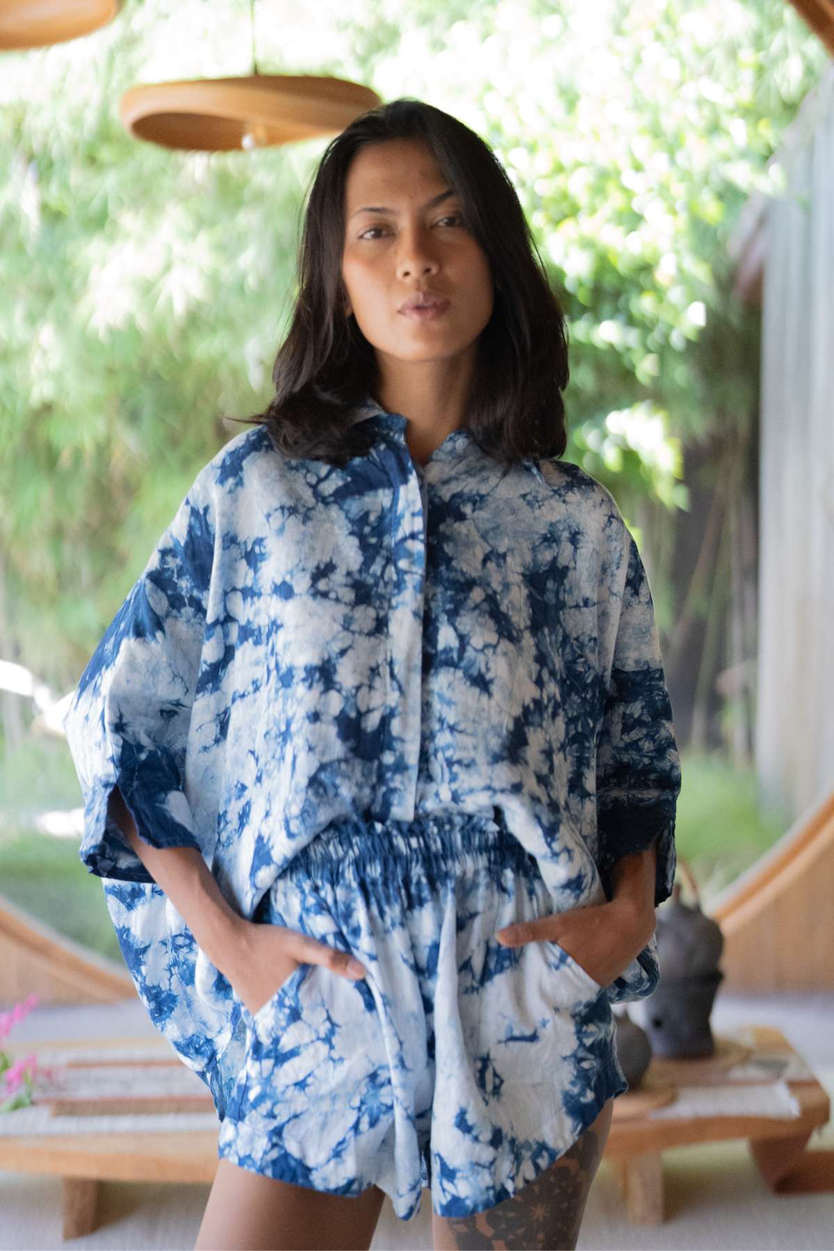 A person confidently stands with hands in pockets, wearing the Indigo Suka Set by Myrah Penaloza, a blue and white tie-dye outfit made from 100% linen and ethically sourced materials. The lush, green garden is visible through large windows in the background.