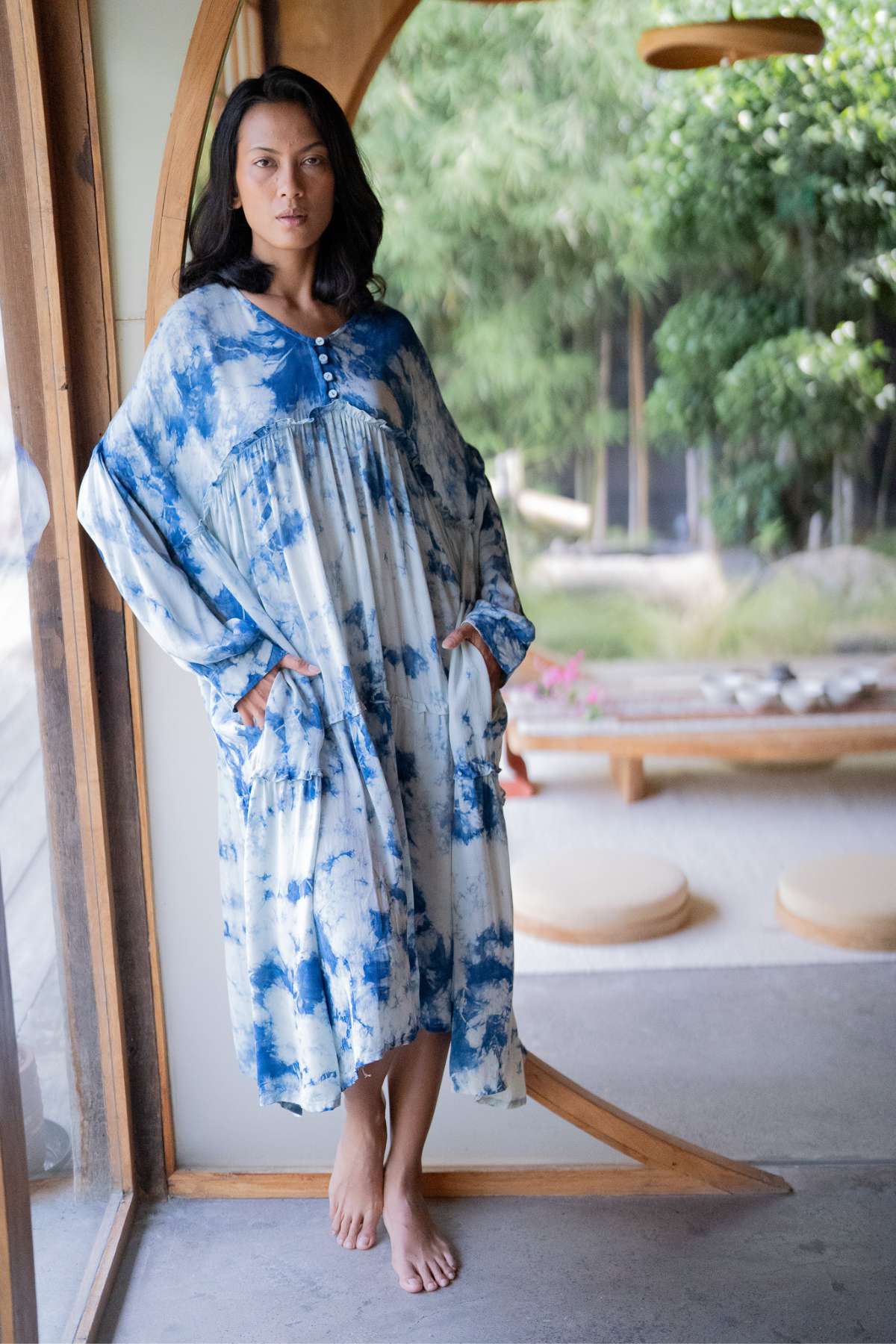 A person stands indoors by a window, adorned in the Indigo Eva Gown made of 100% silk by Myrah Penaloza. With long dark hair, they lean against a wooden frame while green foliage and sleek wooden seating adorn the background.