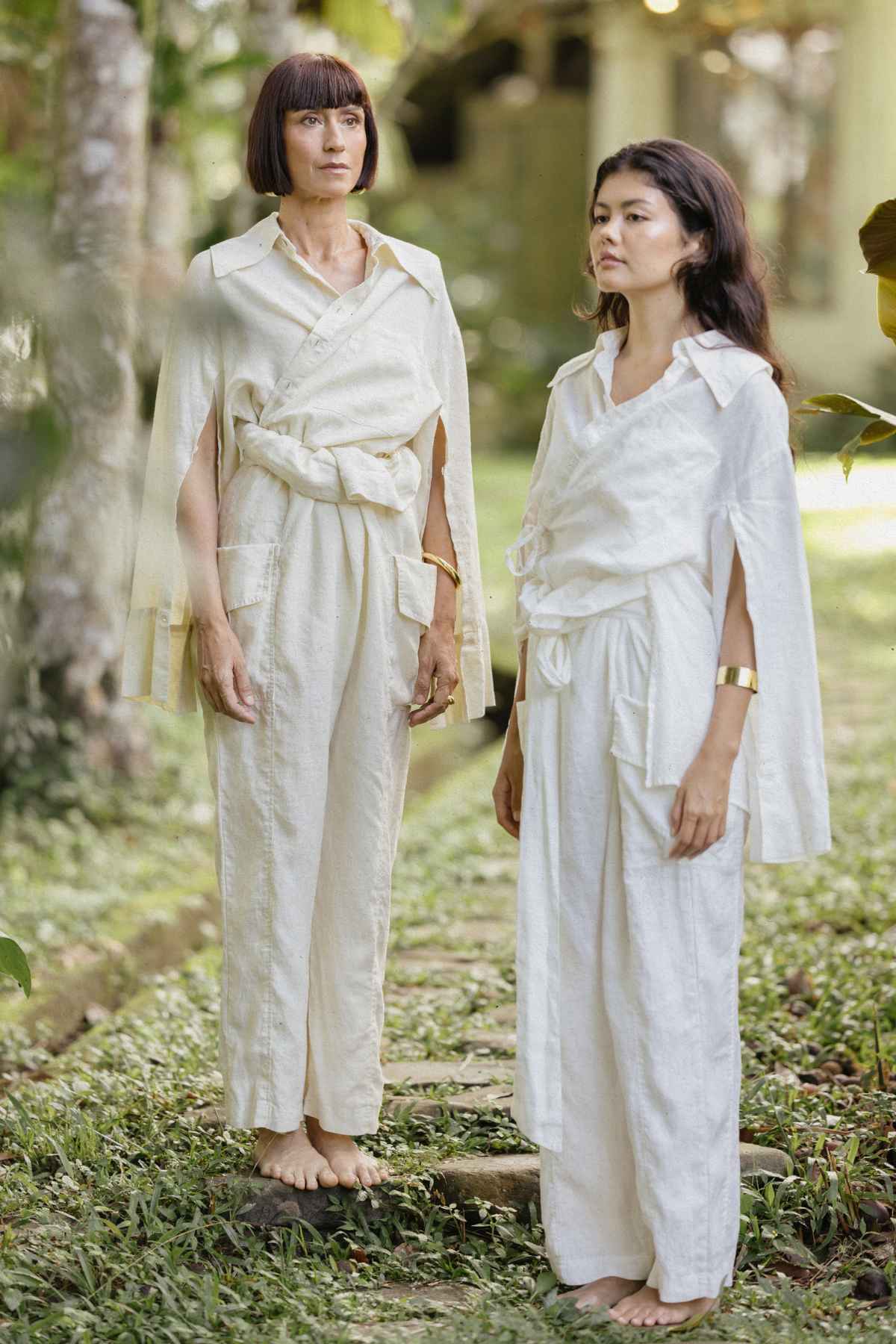 Two barefoot women stand outdoors on a grassy path, their loose-fitting, off-white and cream-colored Sutra Linen Sets from Myrah Penaloza showcasing unique draped designs. This versatile linen set complements the natural, serene environment, with greenery and blurred trees in the background promoting a sustainable lifestyle.