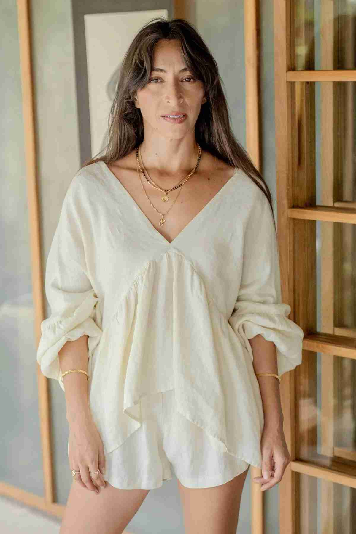 A woman with long, dark hair stands indoors near a wooden window, dressed in the Eva Linen Top by Myrah Penaloza, which features flowering sleeves and is paired with shorts. She accessorizes with layered necklaces and bracelets, smiling softly at the camera as light filters in through the window, creating a warm ambiance.