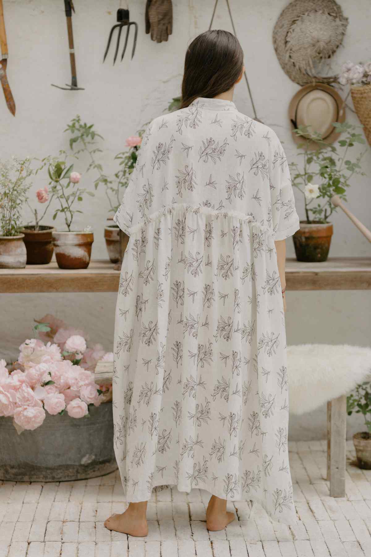 A woman with long brown hair stands barefoot on a tiled floor, facing away from the camera. She is wearing the flowy Bona Dress Cha Dao from Myrah Penaloza's limited edition Tea Collection. Around her are various potted plants, flowers, gardening tools, and hats hanging on the wall.