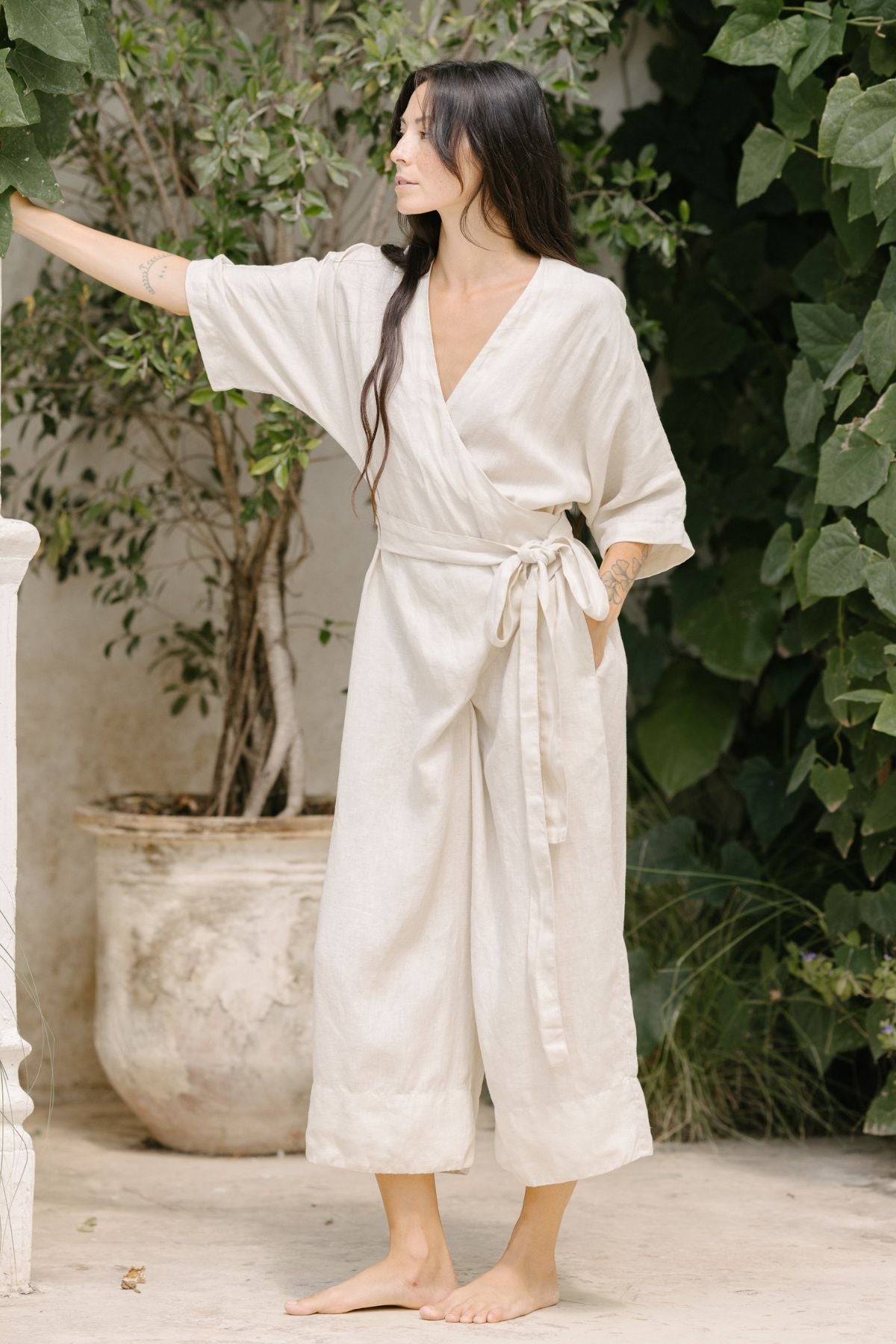 A woman with long dark hair, wearing the Rey Linen Playsuit by Myrah Penaloza—light beige, long-sleeved, and featuring an adjustable front wrap—stands barefoot on a patio. She has one arm resting on a pillar and is surrounded by lush greenery. The scene appears serene and elegant, with soft natural lighting.