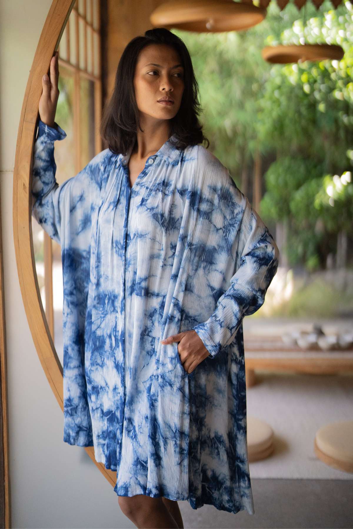 A woman stands by a circular wooden door, wearing the Indigo Dreams Kundalini Gown Short by Myrah Penaloza. She gazes to the side, with one hand on the door. The background reveals a tranquil interior with soft lighting and greenery outside.