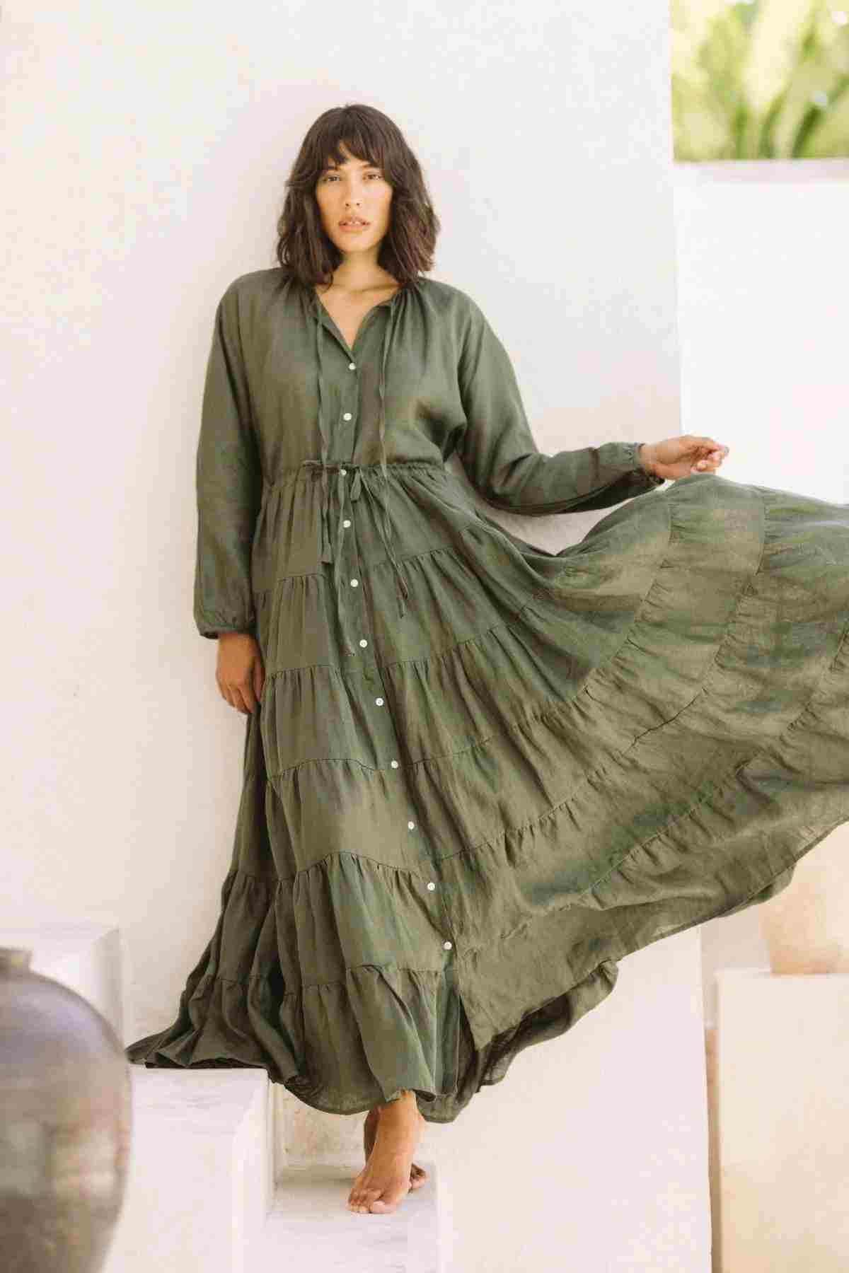 A woman in a flowing green linen La Mexicana Gown by Myrah Penaloza stands on white steps, with one arm extended, creating movement in the dress. The adjustable waist of the gown enhances her silhouette amid the minimalist, light-colored background.