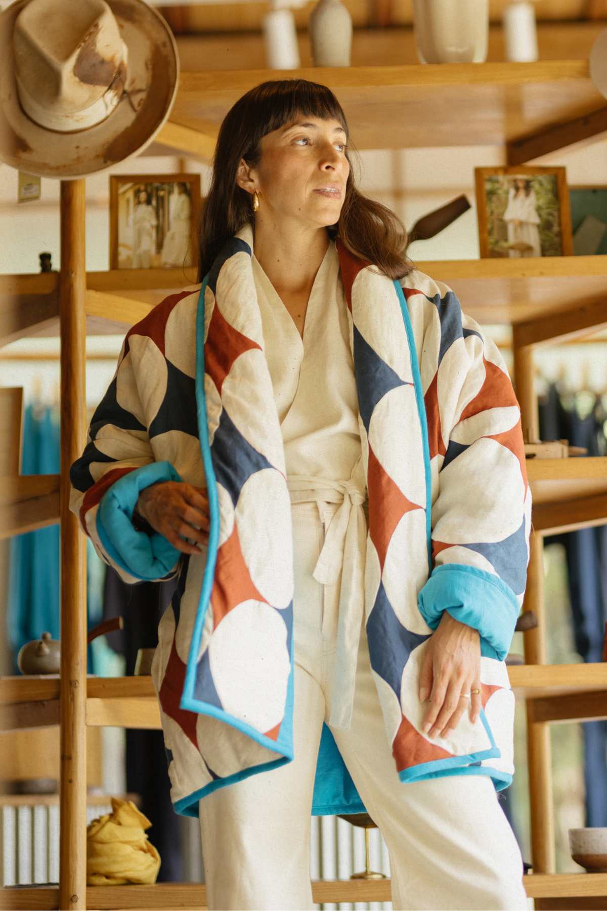 A woman stands indoors in front of wooden shelves, wearing a cream-colored outfit and a Javi Quilted Jacket (Pre-Order) by Myrah Penaloza, featuring a colorful geometric pattern. She gazes slightly upward to her left. The shelves behind her display various ceramics and framed photos.