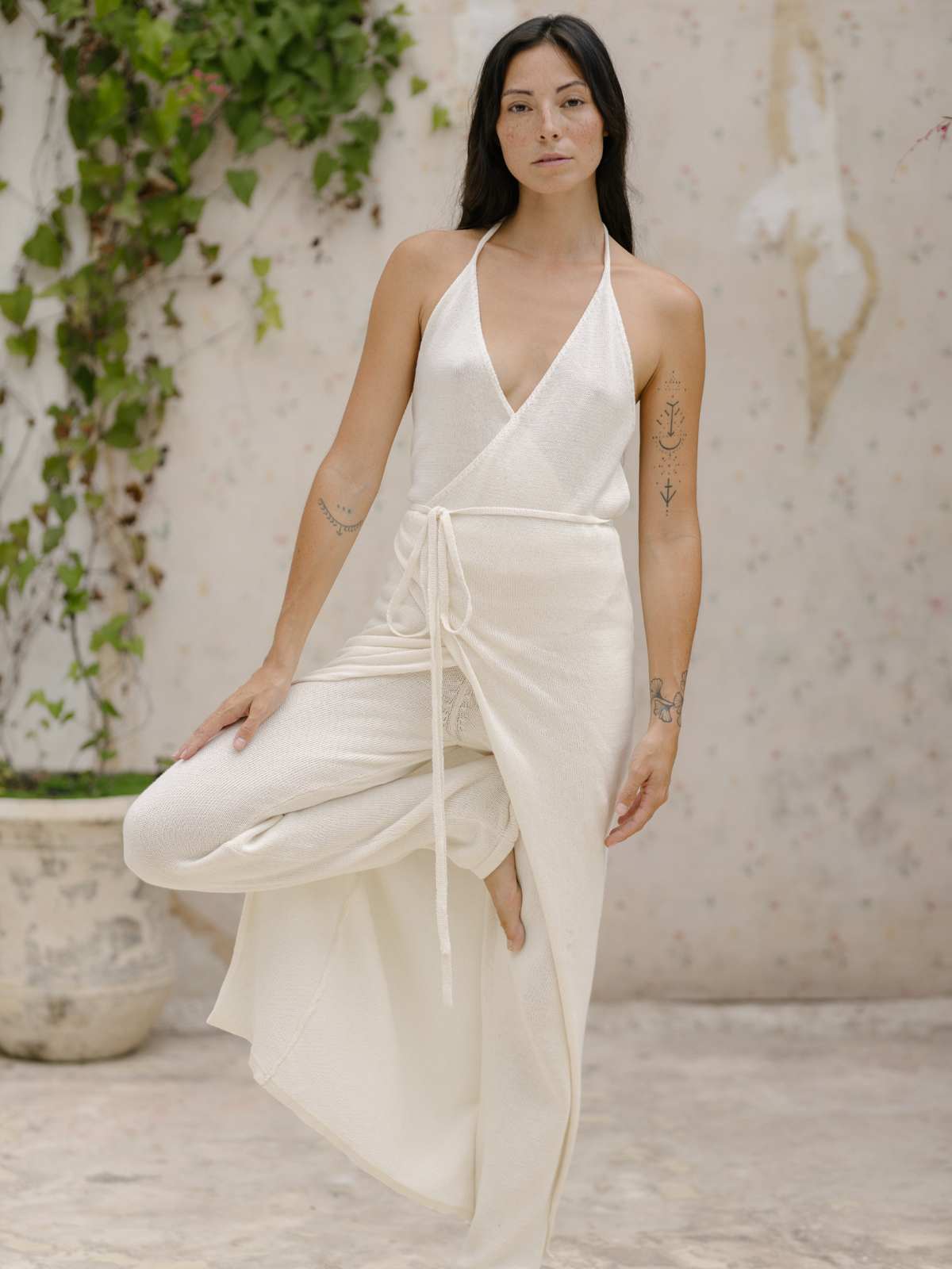 A woman wearing a Honey Cotton Knit Wrap Dress by Myrah Penaloza stands in a yoga tree pose near a wall adorned with vines. She has long black hair and tattoos on her arms, and her expression is serene. The setting is calm and minimalist.