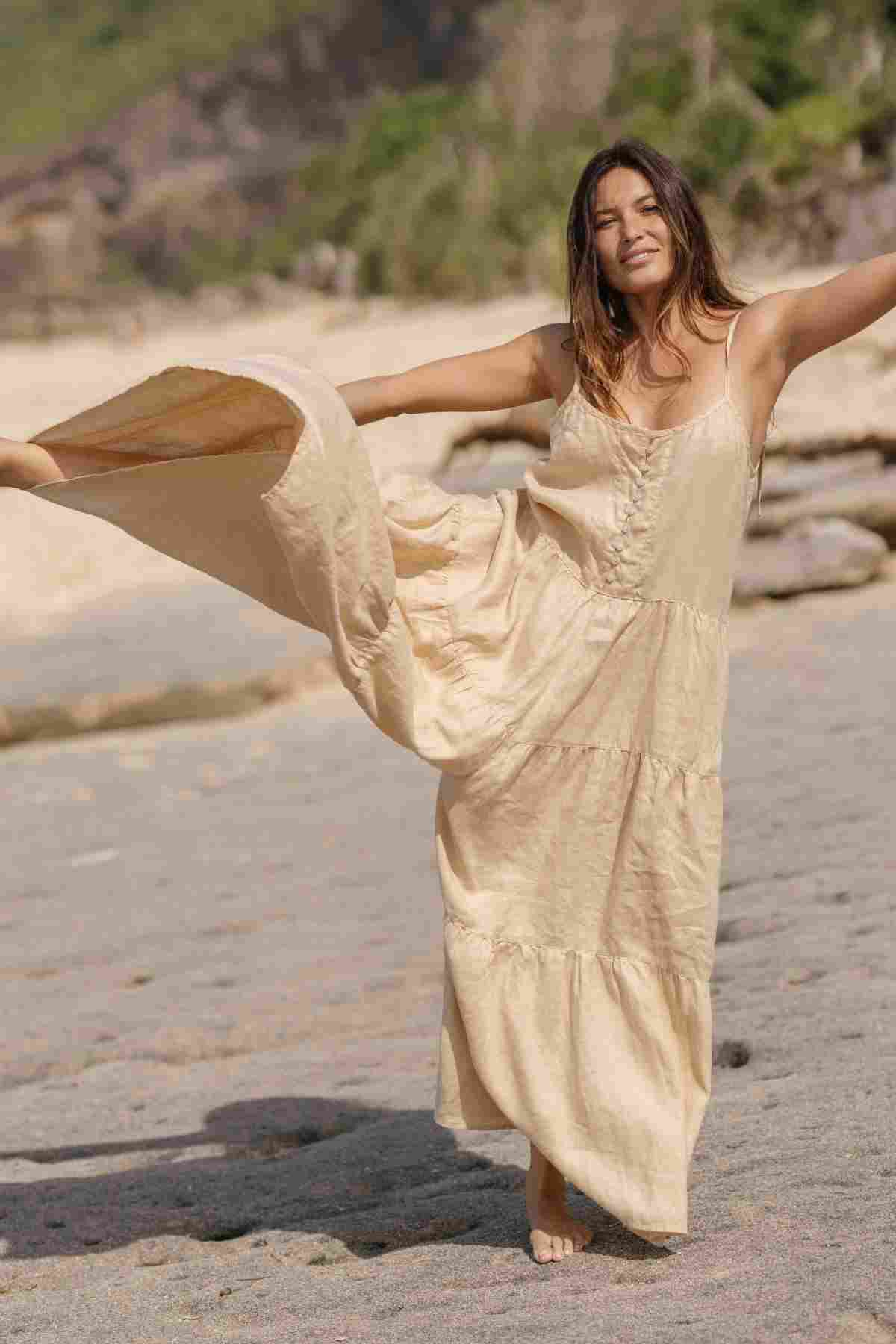 A woman merrily twirls on a beach, wearing the La Mexicana Playsuit by Myrah Penaloza. Her graceful pose with one leg lifted mirrors her joy amidst the backdrop of sand, rocks, and greenery. The playsuit's beige hue and adjustable straps perfectly capture the essence of summer attire. With long hair flowing and a radiant smile, she basks in the sunshine.