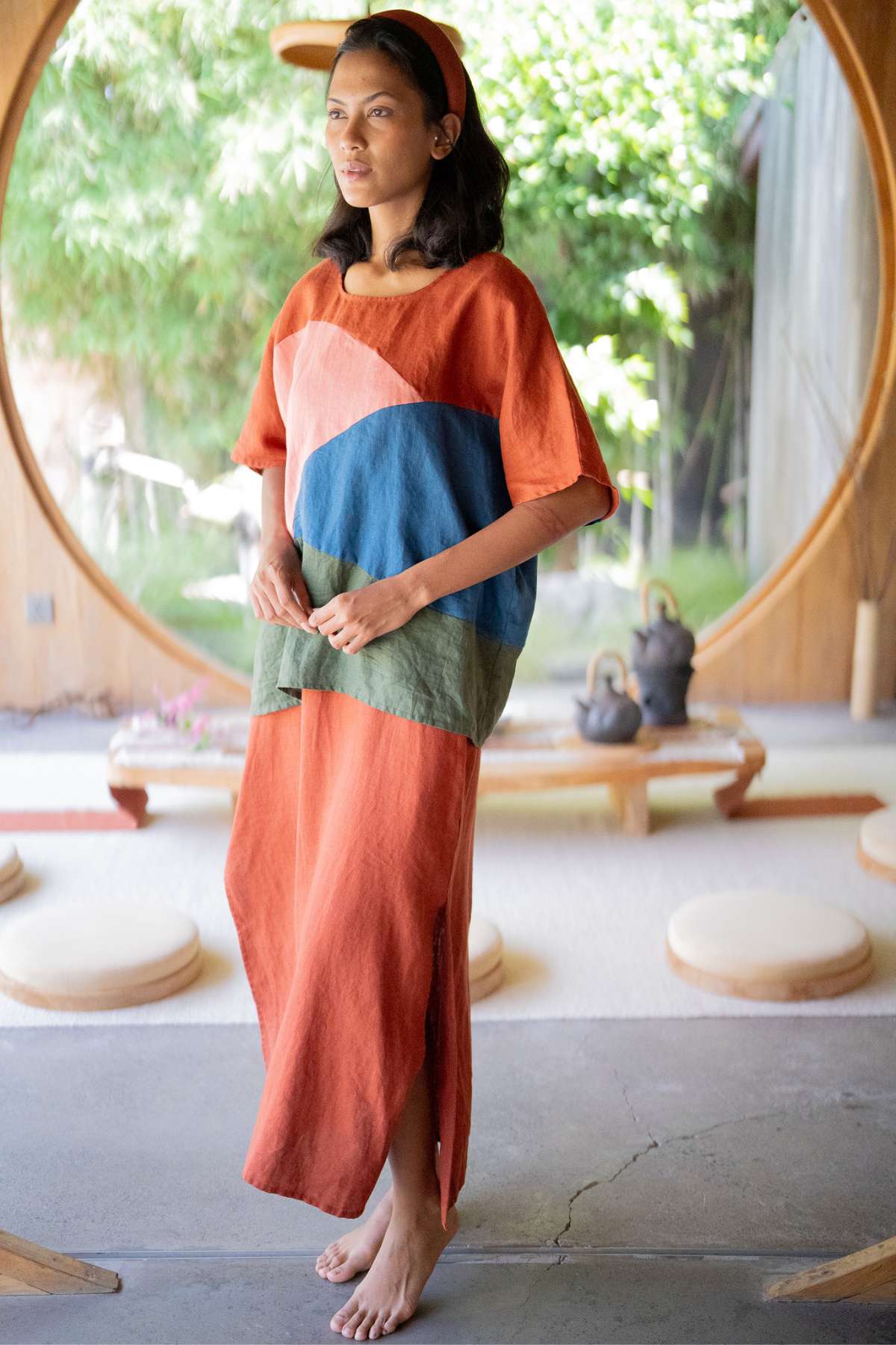 A person stands barefoot in a room with a large circular window, wearing the Uluwatu Top by Myrah Penaloza, adorned with colorful geometric patterns. Surrounded by round cushions and a wooden table topped with teapots, the scene exudes tranquility, framed by the lush greenery outside.