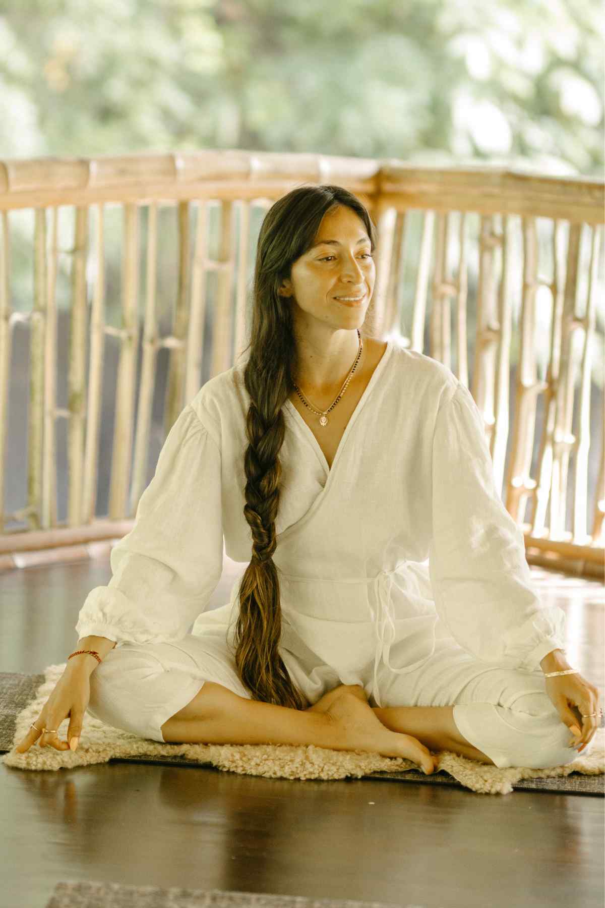 A person with long hair, wearing the Myrah Penaloza Linen Long Sleeve Dora Playsuit, which features unique detailing and an adjustable waistline, is seated cross-legged on a mat in a serene, nature-filled setting. They have a relaxed expression and are surrounded by wooden elements and lush greenery in the background.