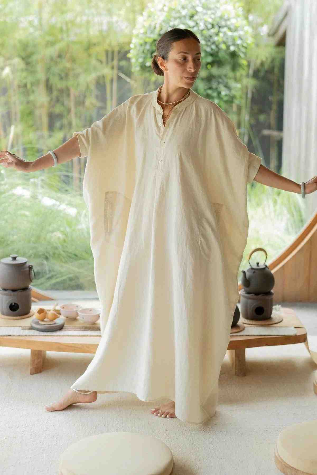 A person stands indoors with arms slightly outstretched, wearing a flowing La Majia Kaftan from Myrah Penaloza in a soothing, cream-colored linen. Their expression is calm and serene. In the background, there is a low wooden table adorned with tea sets and other small items, and a large circular window with a view of a garden reminiscent of Frida Kahlo's sanctuary.