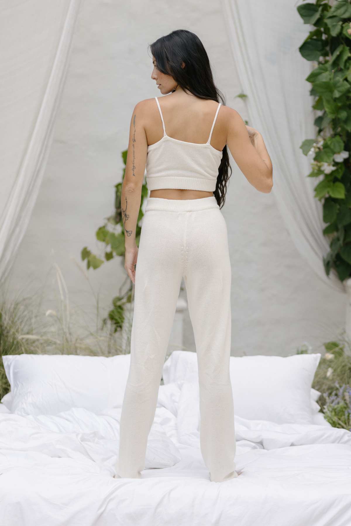 A woman with long dark hair stands on a bed, facing away in her Honey Knit Sets by Myrah Penaloza. The light beige tank top and pants, crafted from natural cotton, blend seamlessly with the outdoor setting adorned with white drapes, green foliage, and cozy pillows and bedding.