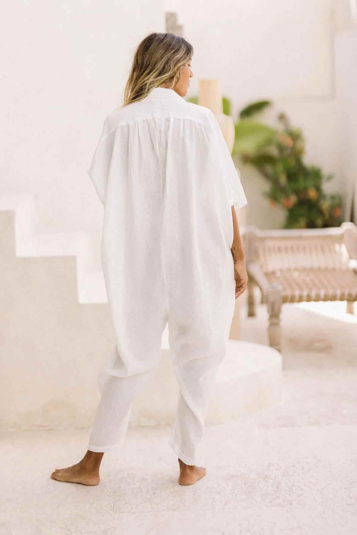 A woman stands barefoot on a light-colored, sunlit floor, facing away from the camera. She is wearing a loose-fitting, white Linen Sat Torri Playsuit with wide dolman sleeves by Myrah Penaloza, and her hair is down. A staircase and some furniture are visible in the background, along with green plants. The scene exudes a retro 1980s inspired design.