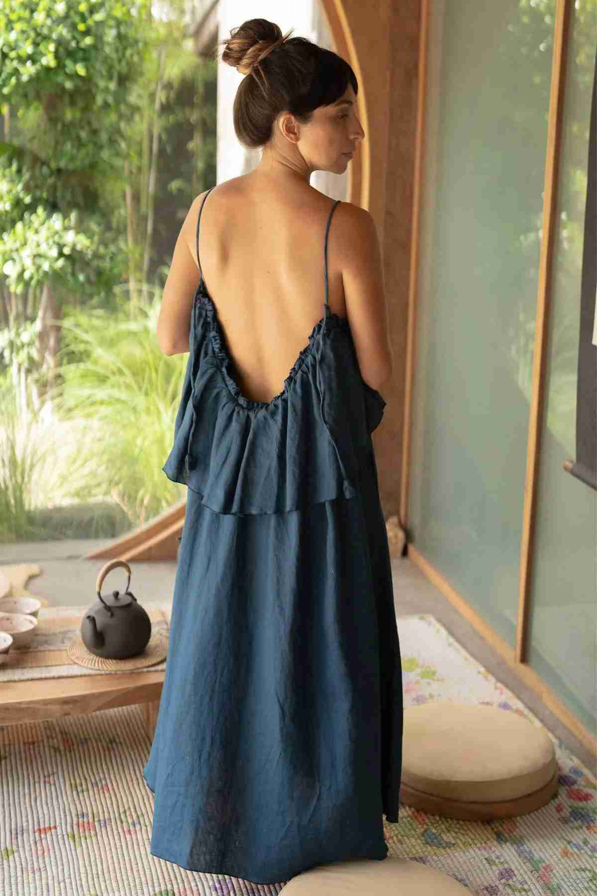 A woman stands with her back to the camera, wearing the Linen Muse Gown by Myrah Penaloza, a dark blue stone-washed linen dress featuring adjustable straps and ruffled details. Her hair is tied in a bun. She is on a porch adorned with a wooden bench, round cushions, a woven rug, and a tea set on a tray. Lush greenery can be seen outside.