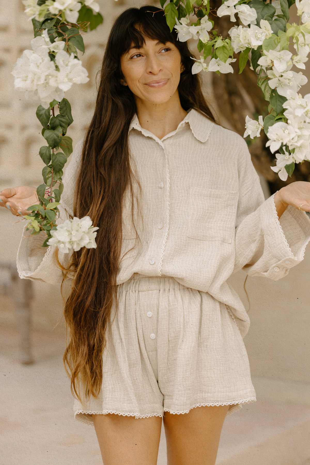 A person with long hair smiles beneath a branch of white flowers while wearing the Nidra Button Down Set Lace Edition by Myrah Penaloza. Their light-colored, loose-fitting shirt and shorts showcase sustainable beauty, with a softly blurred background suggesting an idyllic outdoor setting.