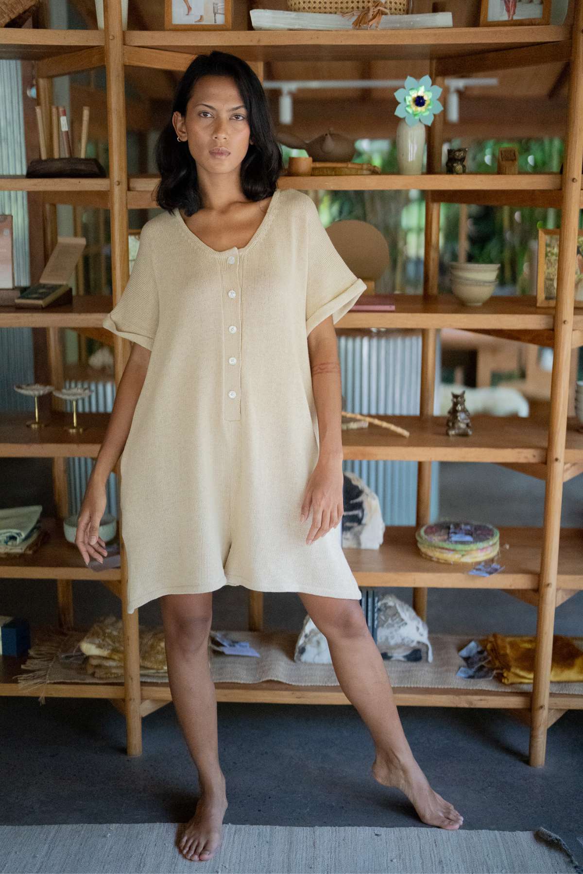 A person stands barefoot indoors, wearing the Henley Knitted Playsuit - BF by Myrah Penaloza, which is subtly breastfeeding friendly. The background features wooden shelves with various decorative items, creating a cozy and relaxed atmosphere.