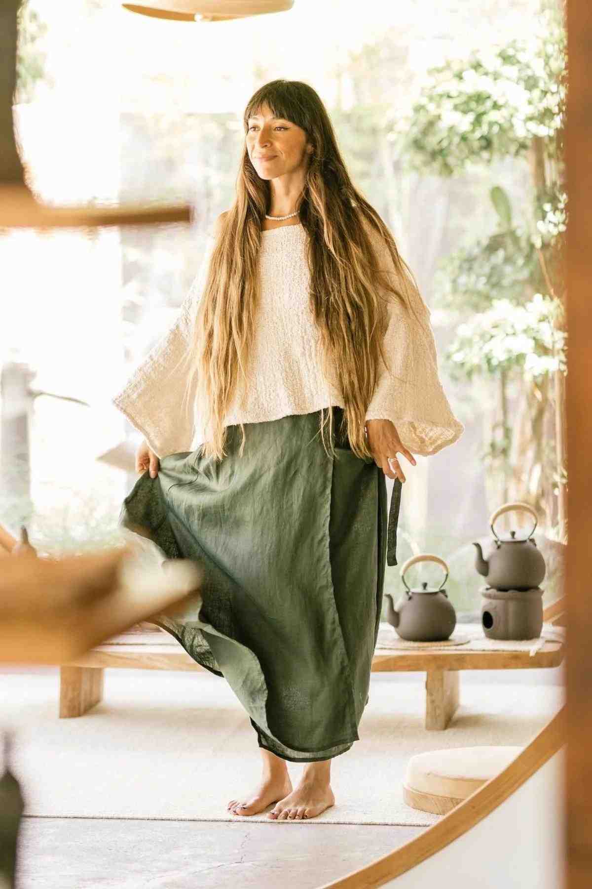 A woman with long hair stands barefoot indoors, adorned in the Spirit Top & Shanti Wrap Skirt Set by Myrah Penaloza. The loose, white top pairs beautifully with the adjustable green skirt made from Hand-Woven Tummangal Cotton. She exudes a calm, content expression as she gracefully holds the skirt. In the background, teapots and a serene, sunlit space with plants and wooden elements create a tranquil ambiance.