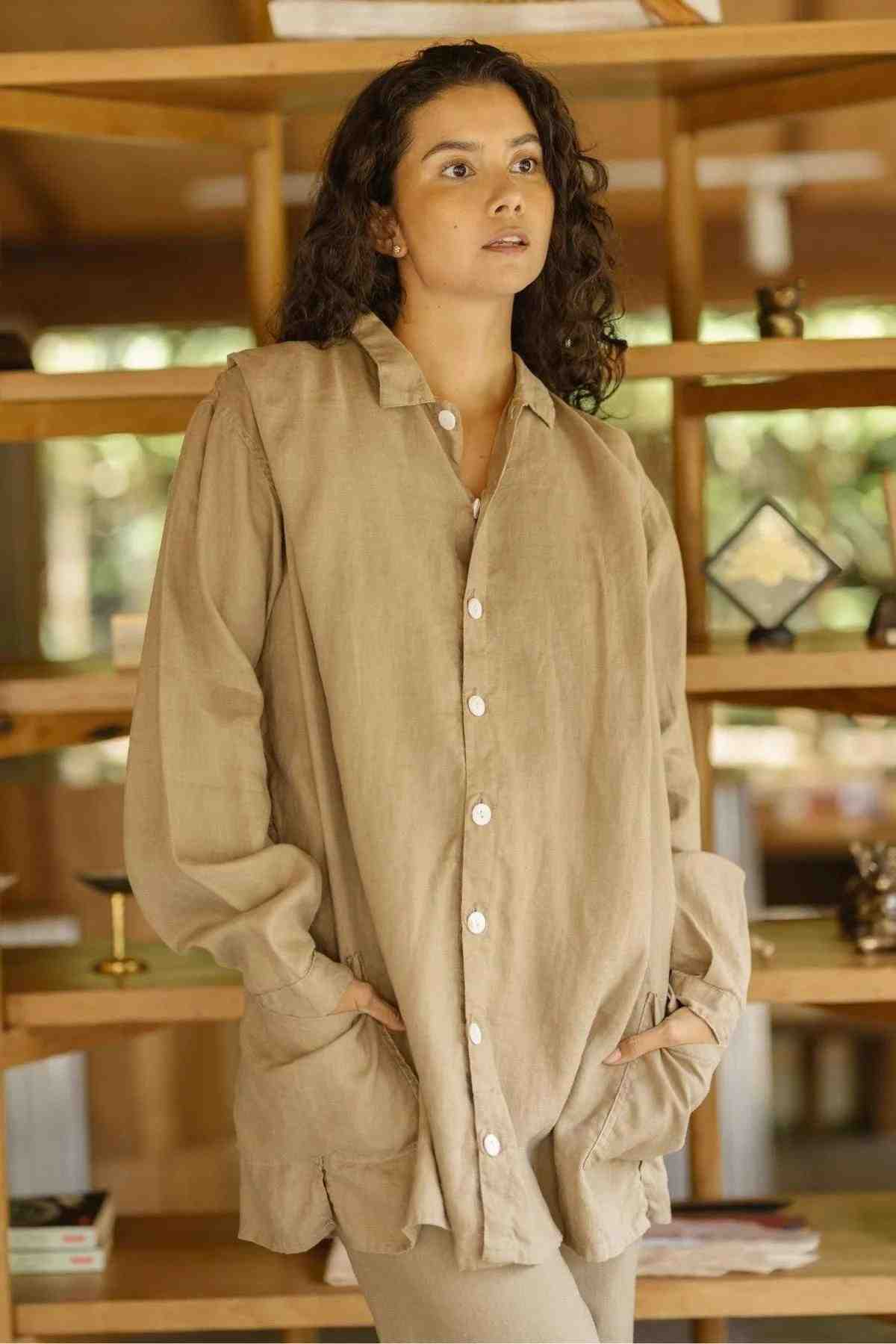 A person with long, curly hair stands in a relaxed pose, wearing a loose, casual beige button-up shirt paired with the Tender Jacket (100% Linen) in deep royal blue by Myrah Penaloza, their hands in their pockets. The background features wooden shelves filled with various decorative items and a softly lit indoor setting.