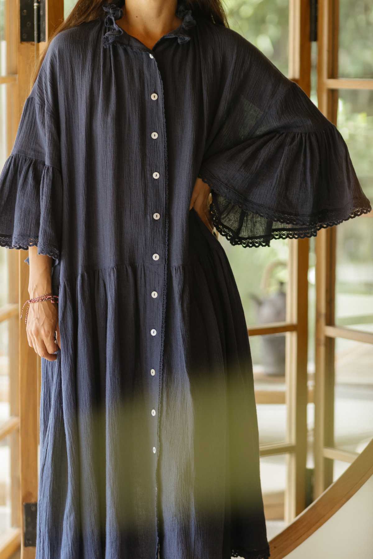 A person stands with a hand on their hip, wearing Myrah Penaloza's exquisite Juliette Linen Gown, characterized by its flowing navy blue fabric, large sleeves, and button-down front. In the background, a wooden-framed window beautifully combines indoor and outdoor elements to elevate the sophistication of this bespoke creation.