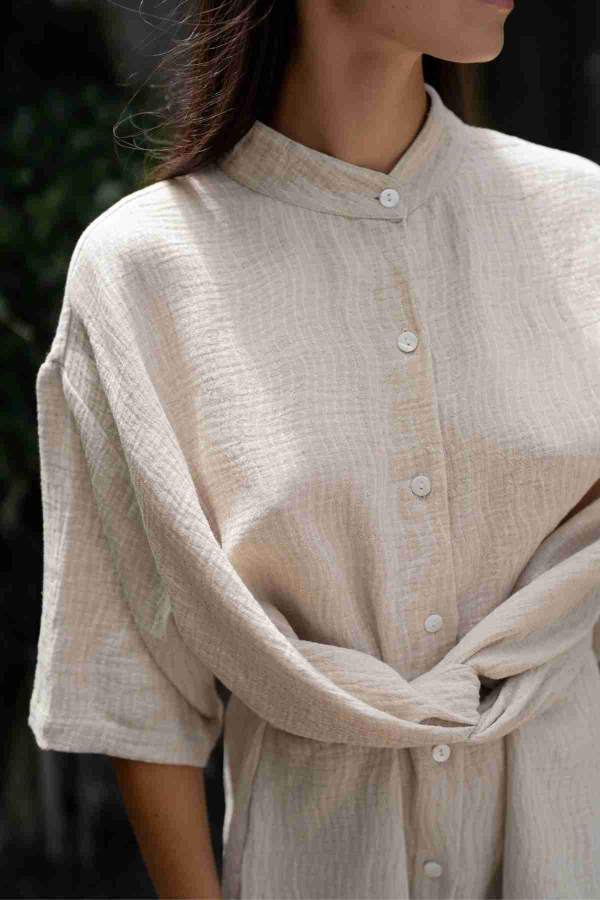 A person is wearing the Pine Playsuit from Myrah Penaloza, a beige, textured button-up shirt made from a cotton linen blend, tied in a knot at the waist. The shirt features mid-length sleeves and a mandarin collar. The person is looking slightly to the side, with their face not fully visible. The background showcases blurred greenery.