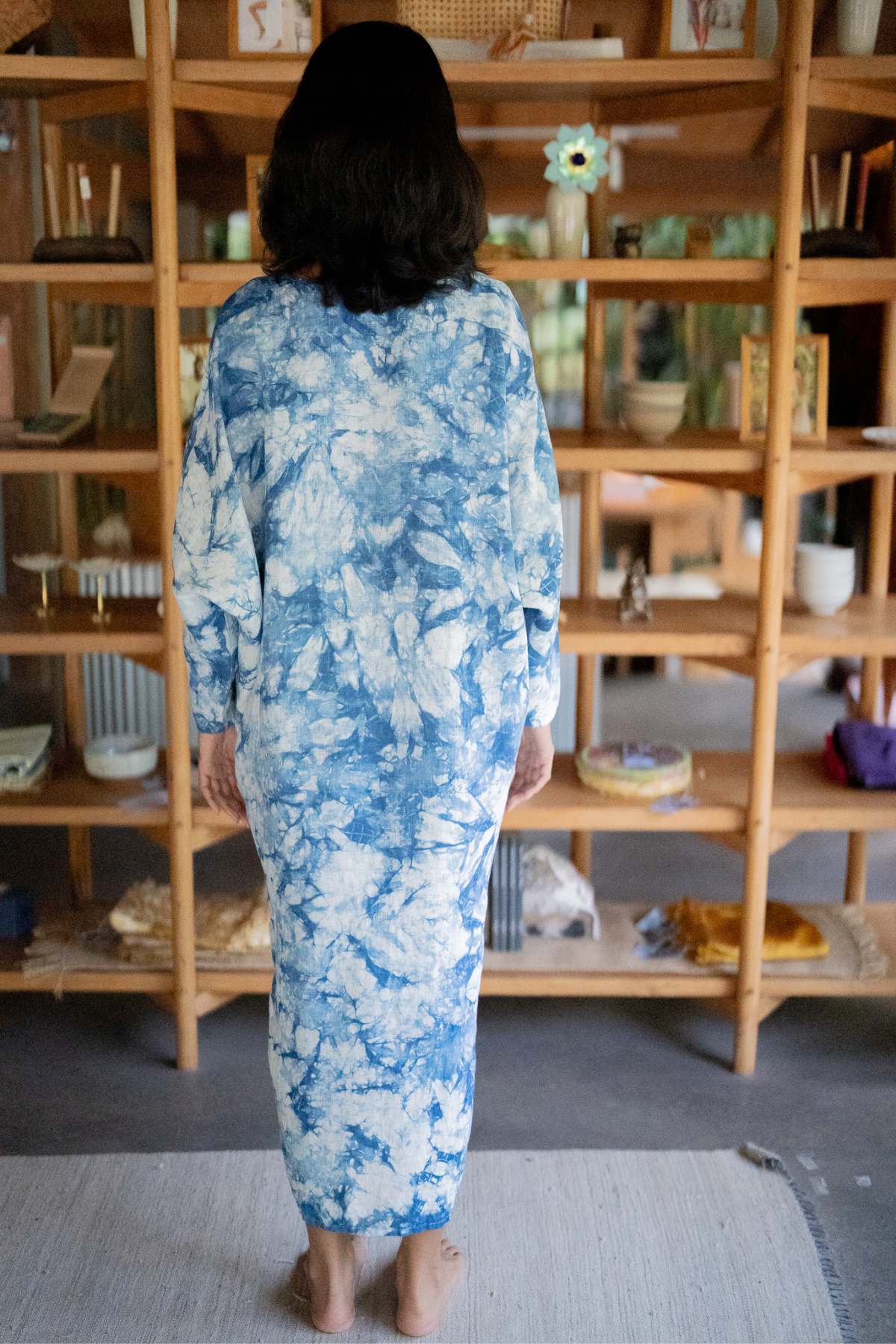 A person with shoulder-length hair stands barefoot, facing a wooden shelf filled with various items. They are wearing the Indigo Virgo Rising Kaftan (One of a Kind) from Myrah Penaloza, made of 100% linen that flows elegantly. The room exudes a cozy and eclectic decor.