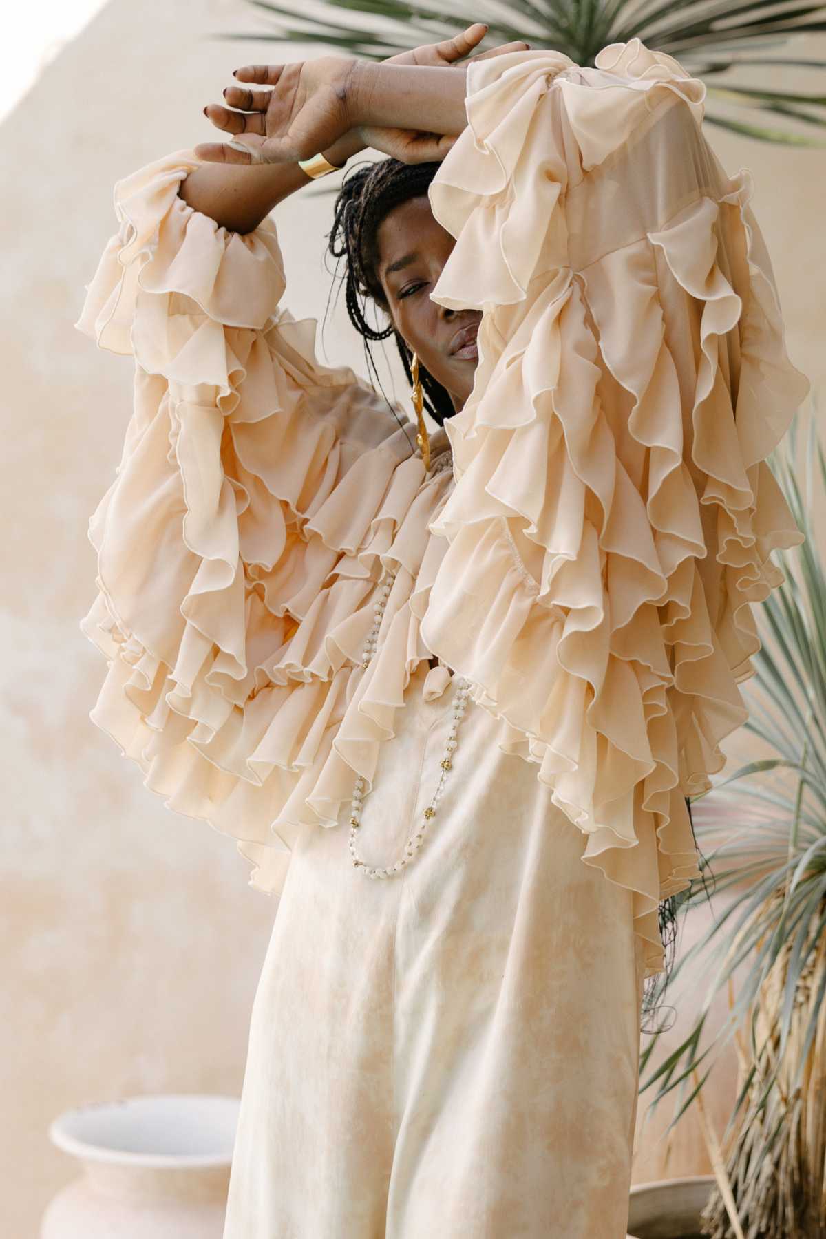 A person poses gracefully with eyes closed, wearing the Anusara Bloom - Made To Order top from Myrah Penaloza. The flowing, ruffled light pastel peach top beautifully complements their Chiffon Sheer Silk dress. They have long braided hair and are standing against a blurred, neutral-toned background with one arm elegantly raised above their head. This nature-inspired couture piece truly exemplifies limited edition elegance.