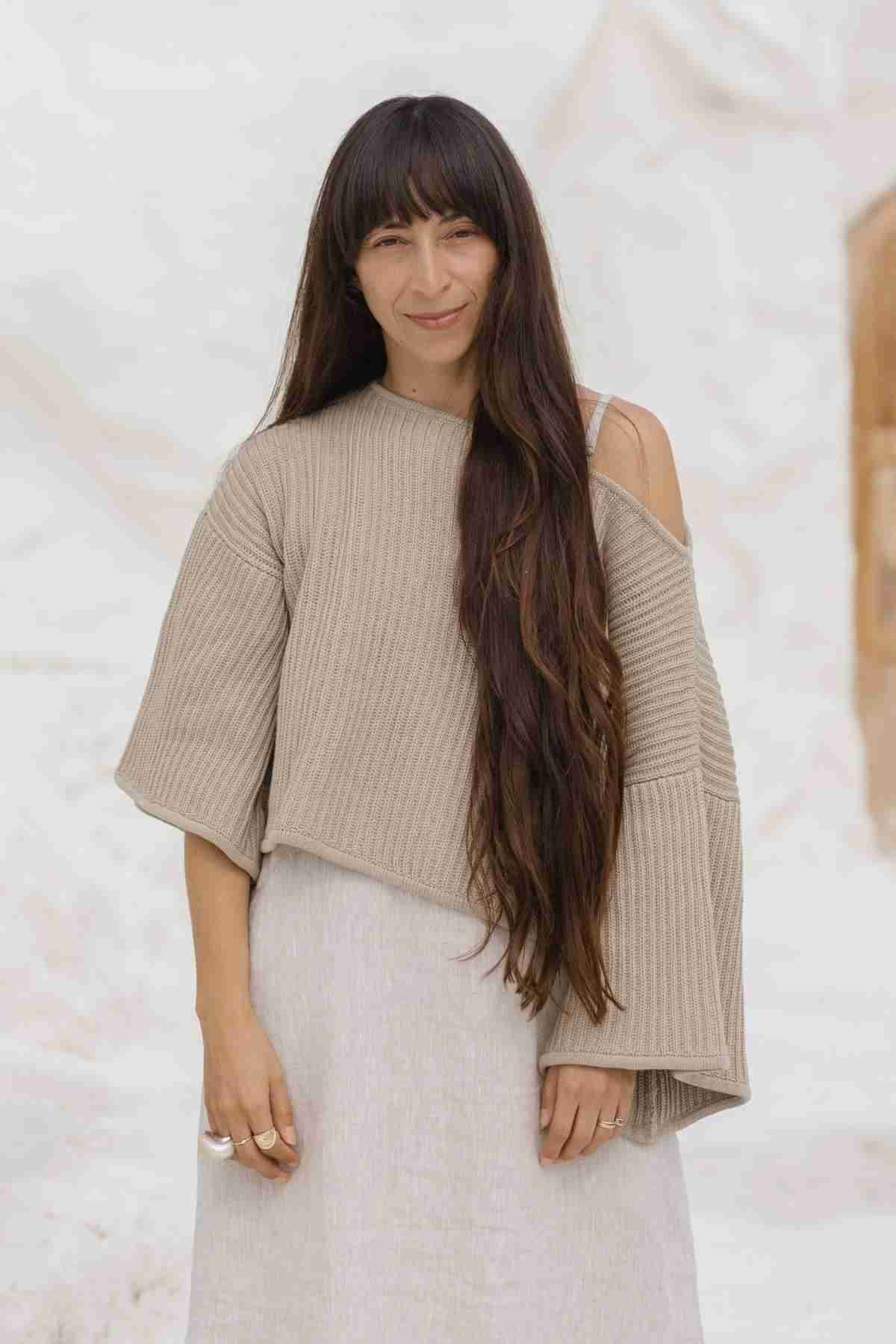 A woman with long, dark hair stands in front of a light-colored background. She is wearing an oversized Spirit Top Sweater Knit by Myrah Penaloza, made from luxurious beige ribbed cotton fabric, with one shoulder exposed and a light-colored skirt. She has a slight smile on her face and her hands are relaxed by her sides.