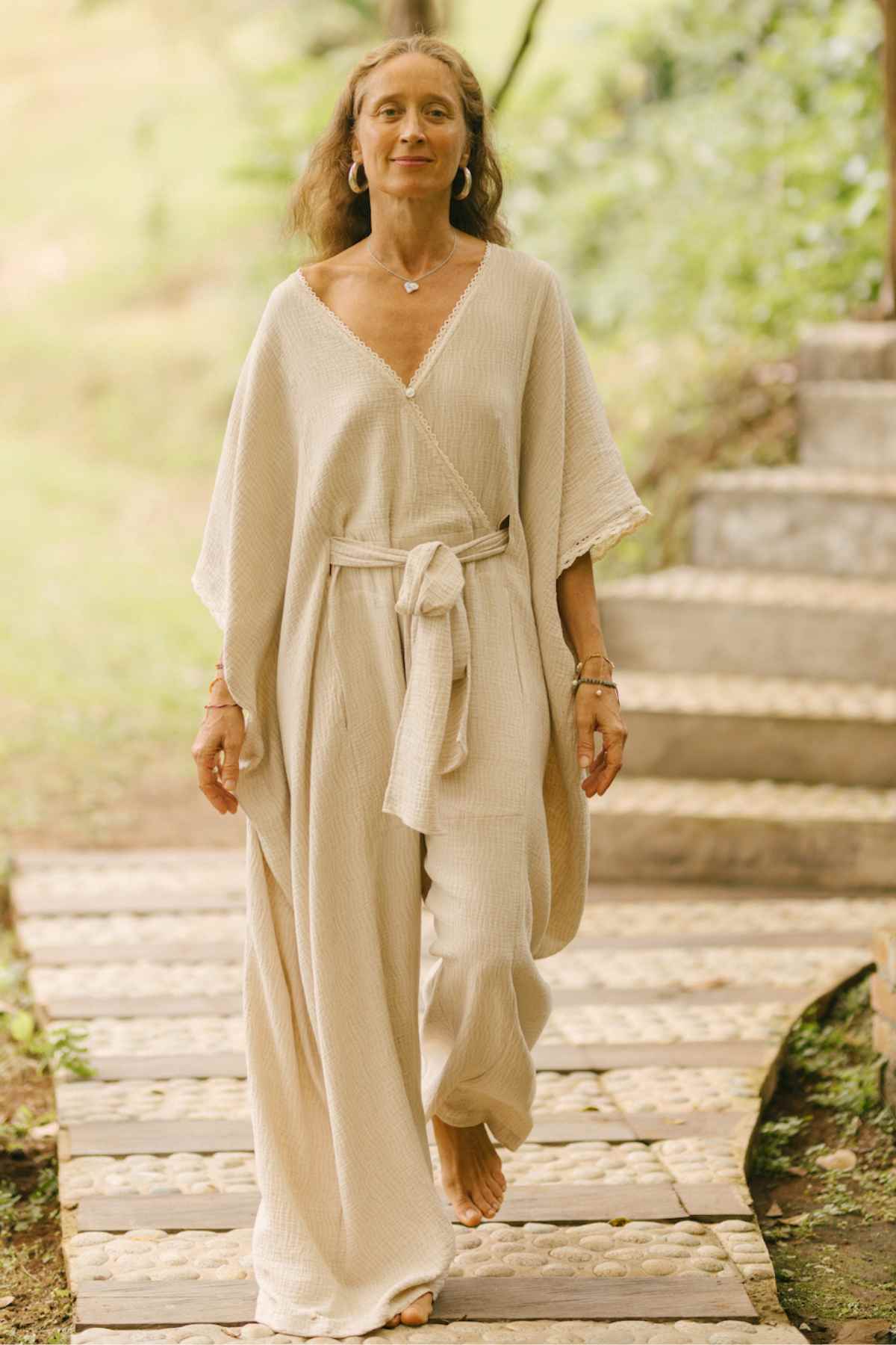 A woman with long hair walks barefoot on a wooden path in a lush, green outdoor setting. She is wearing a flowing, light-colored playsuit with wide sleeves and a belt tied at the waist. Serene and adorned with simple jewelry, she resembles an ethereal vision in her Myrah Penaloza Limited Edition Hand Embroidered River Playsuit.