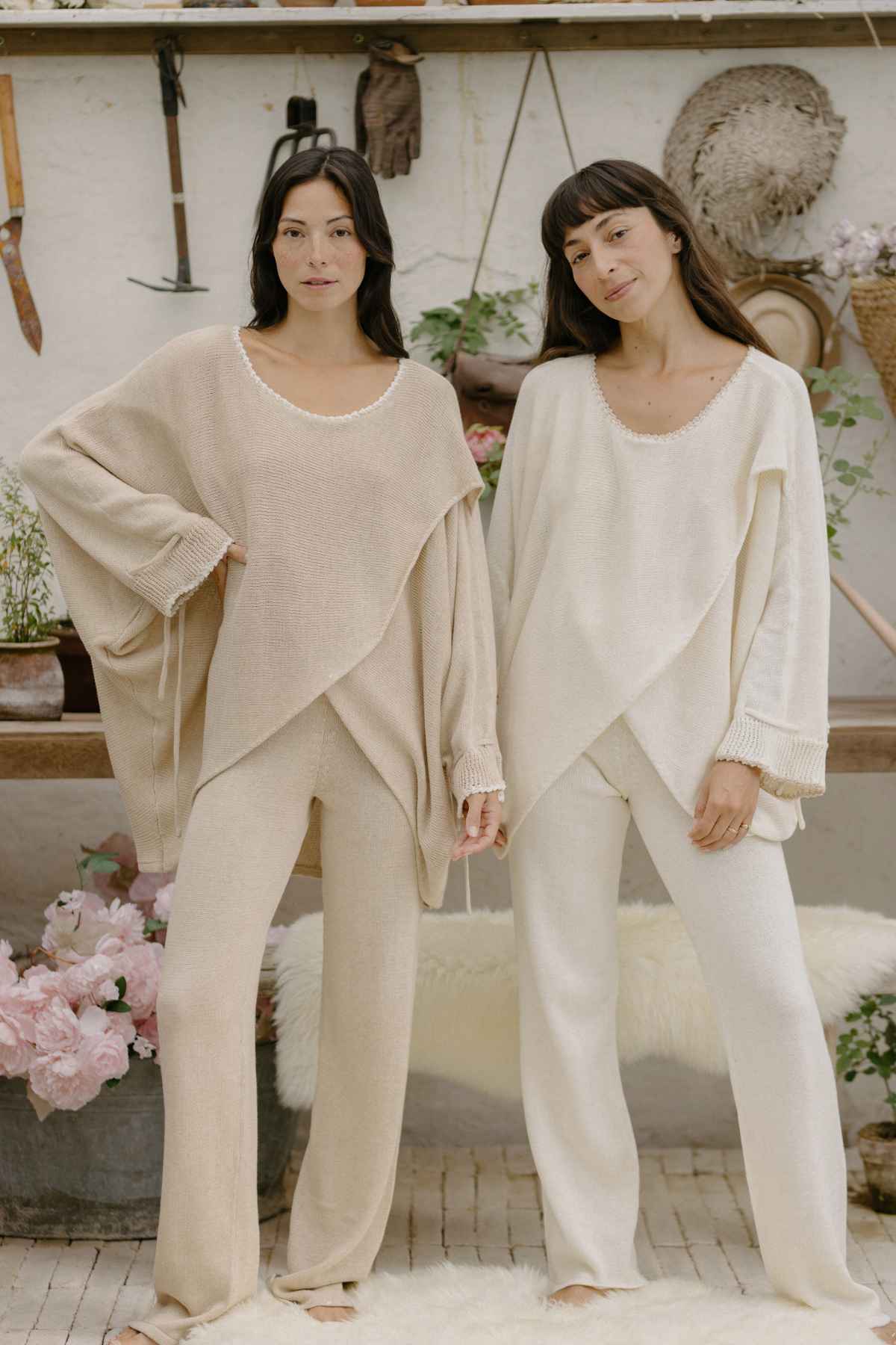 Two women stand side by side in a cozy, rustic setting. Both wear the Honey Knit Sweater from Myrah Penaloza with matching wide-legged pants; one in beige and the other in off-white. Behind them are flowers, plants, a wooden shelf, and various hanging gardening tools.