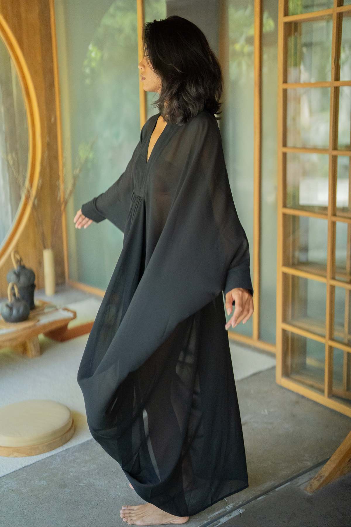 Barefoot and serene, a woman graces the setting adorned with wood and glass elements, like a round window and sliding door. Draped in the flowing Myrah Penaloza Leo Kaftan Chiffon Sheer, she exudes an aura of luxurious power, her arms gently outstretched as she stands indoors.