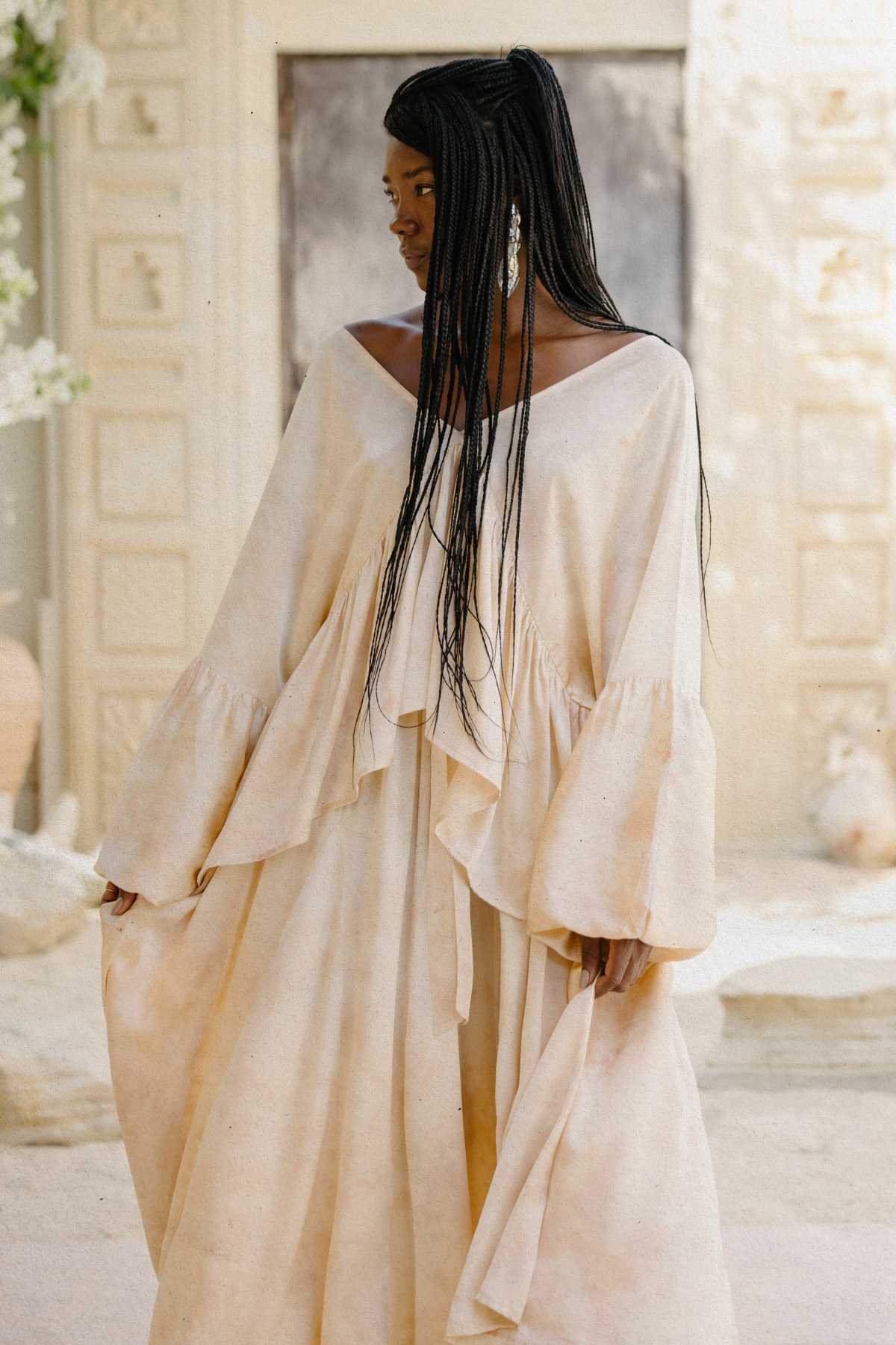 A woman with long braided hair stands gracefully outdoors, wearing the Eva Top & Orisa Dance Skirt Set by Myrah Penaloza. This flowing silk ensemble, botanically hand-dyed in a beige hue and featuring ruffled sleeves, complements the cream-colored wall and ornate door behind her. Her earrings add an elegant touch to her look.