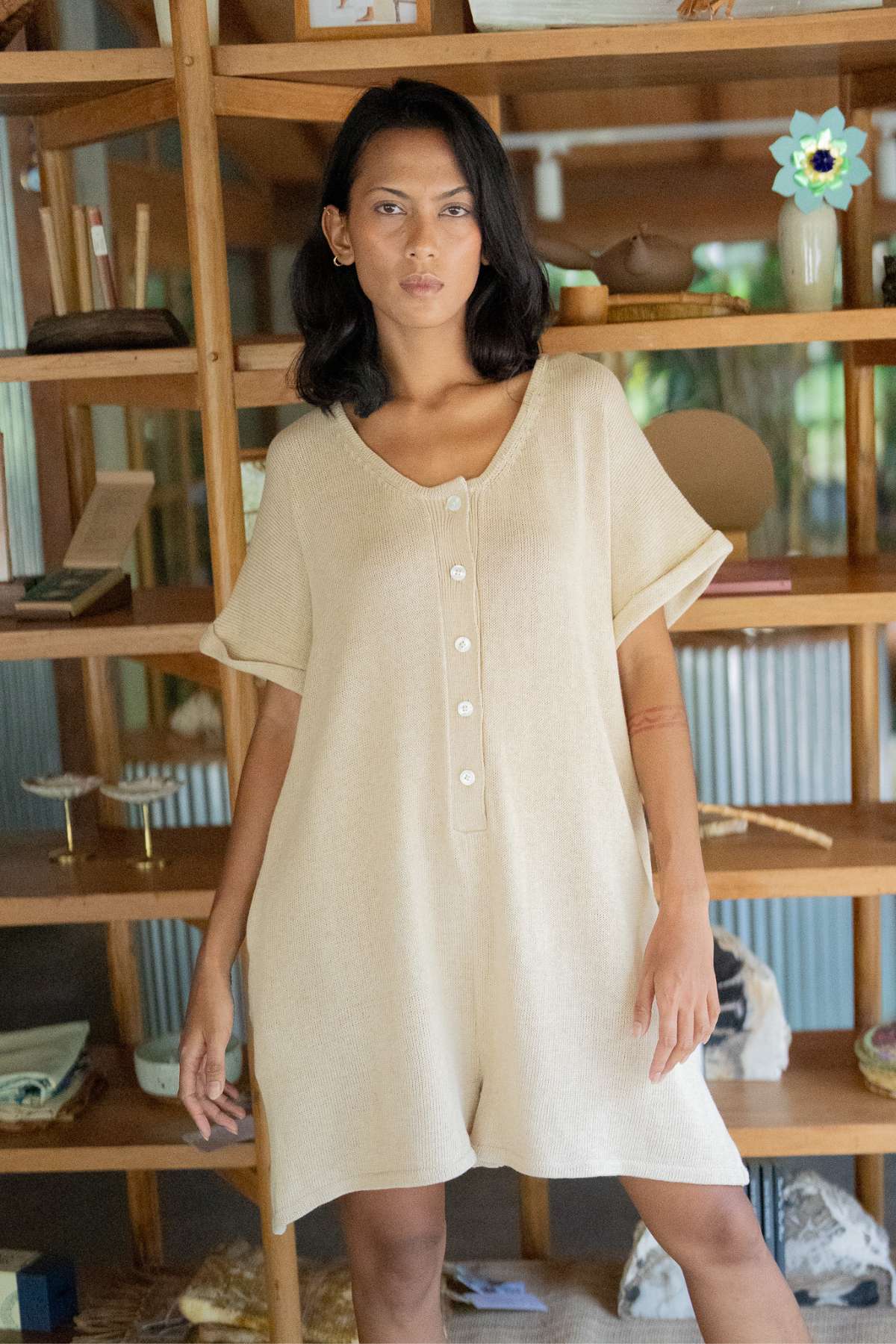 A person wearing the Henley Knitted Playsuit - BF by Myrah Penaloza stands in front of a wooden bookshelf adorned with decorative items and books. A window in the background reveals lush greenery outside, enhancing the serene ambiance.