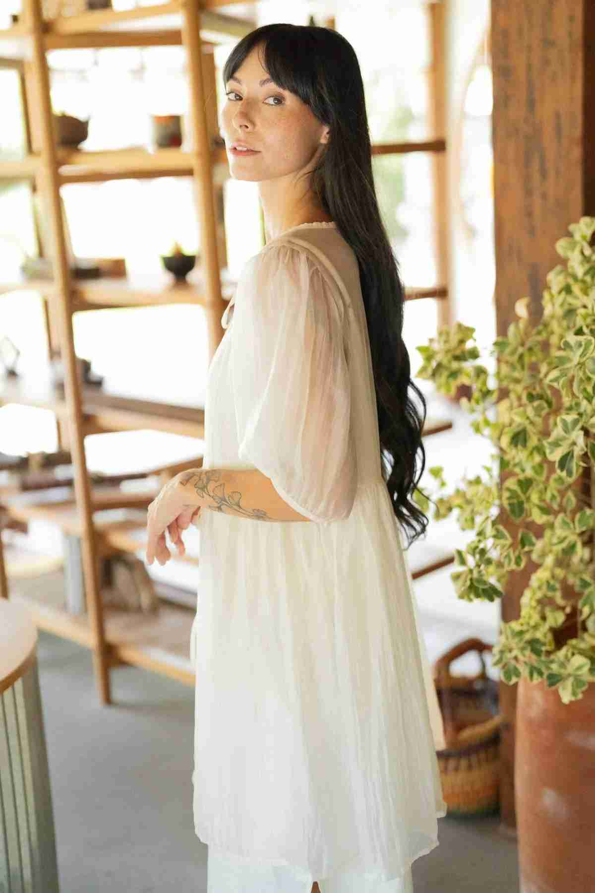 A woman with long black hair and tattoos on her arm is standing indoors, slightly turning towards the camera. She is wearing a Dynasty Sheer Silk Dress by Myrah Penaloza, showcasing its flowy elegance. In the background, there are wooden shelves with various items and a large leafy plant.