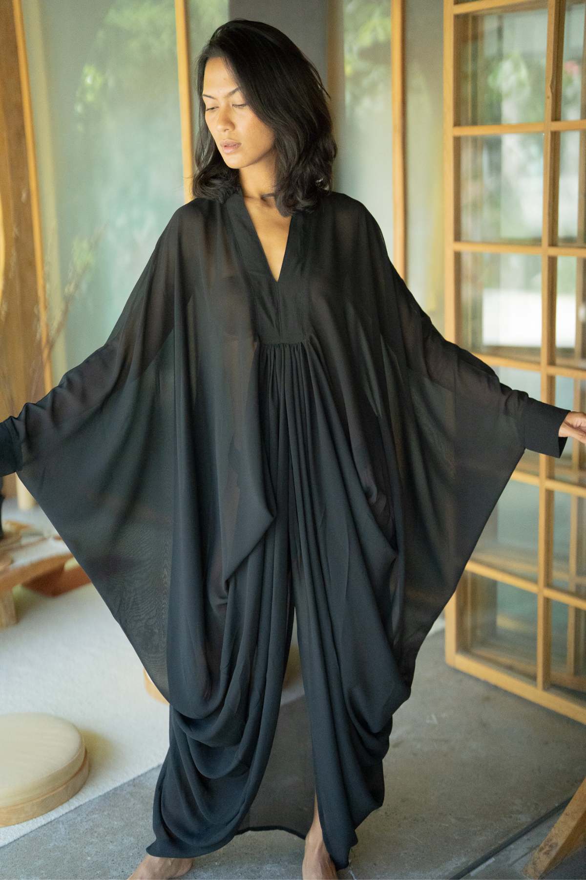 A woman stands indoors wearing the Leo Kaftan Chiffon Sheer by Myrah Penaloza, a flowing black long-sleeved dress that exudes luxurious power. She poses with arms outstretched, highlighting the billowy sheer fabric of the dress. The room features wooden and glass elements, creating a serene ambiance.