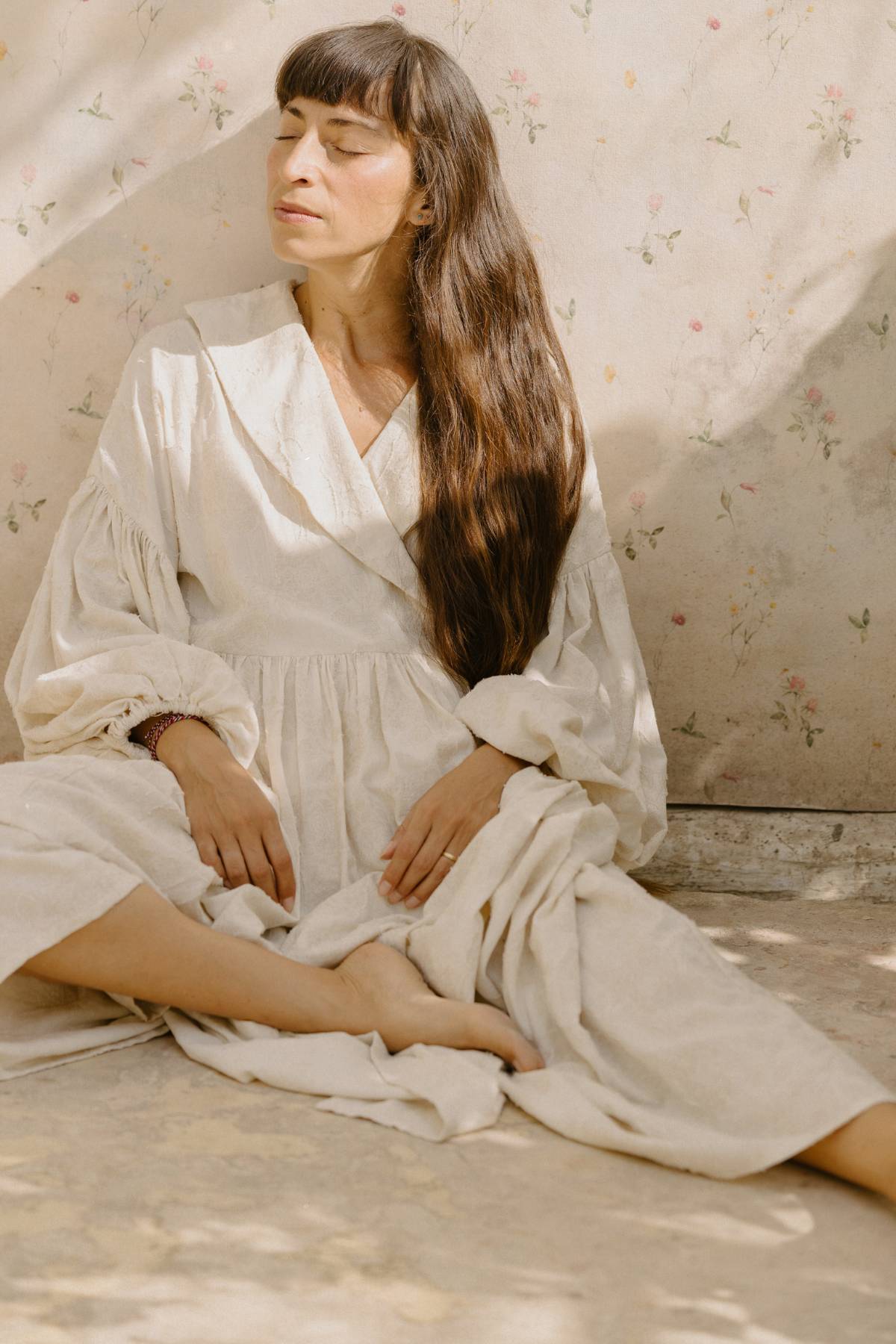 A woman with long dark hair sits cross-legged on the floor, wearing the Blooming Magdalena Dress by Myrah Penaloza. She has her eyes closed and leans against a wall with a floral pattern, while sunlight casts soft shadows around her.