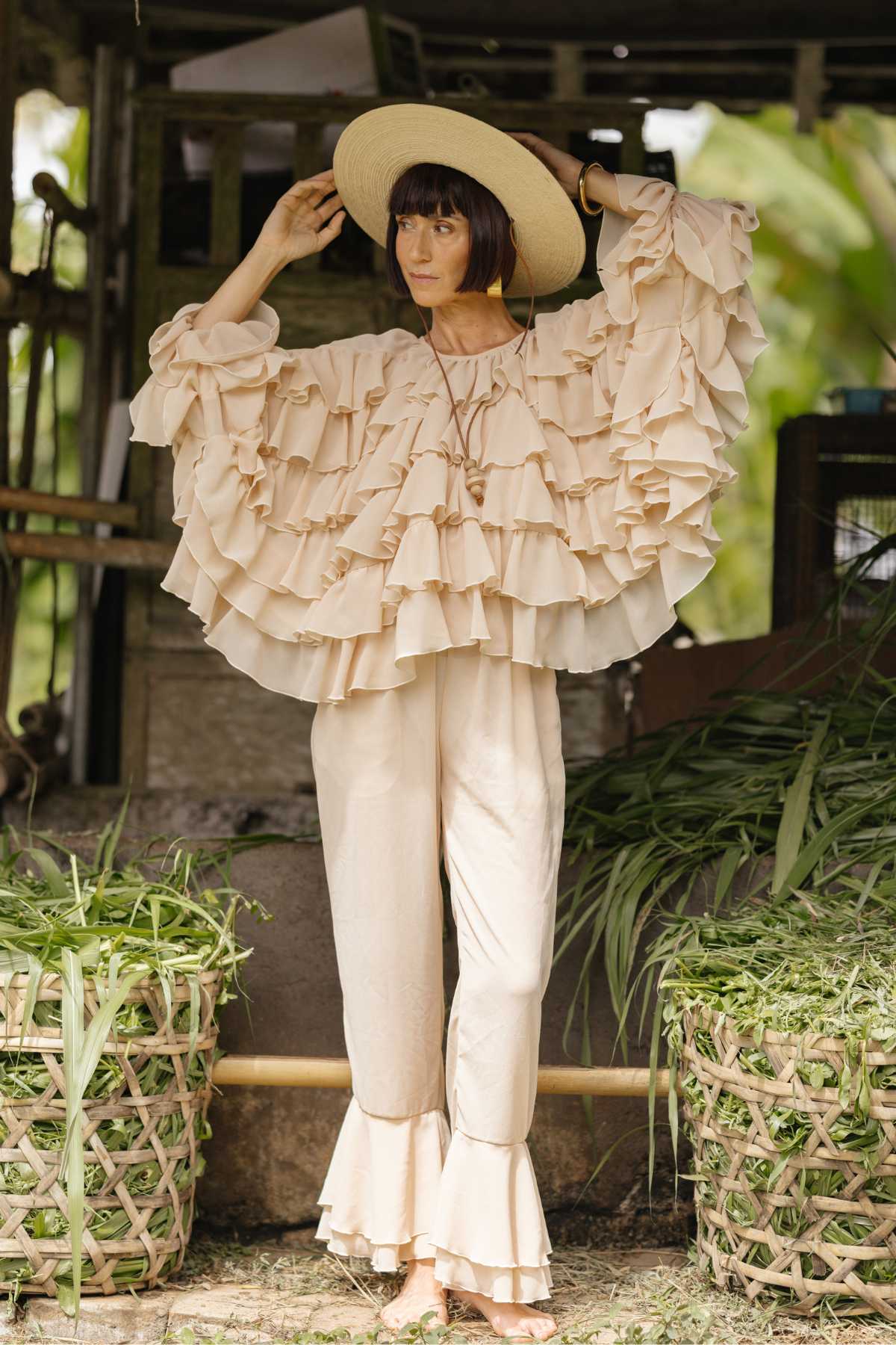 A person wearing the Anusara Bloom - Made To Order ensemble by Myrah Penaloza, featuring a flowy, ruffled beige top and matching chiffon sheer silk pants, stands in front of a rustic setting with baskets of green foliage. They are adjusting a wide-brimmed straw hat while looking off to the side. The background includes wooden structures and greenery.