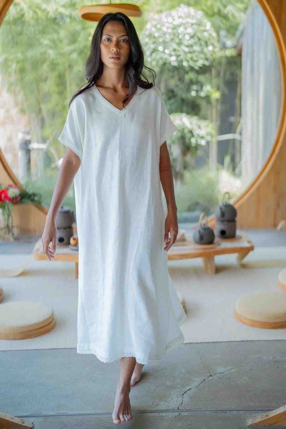 A woman with long dark hair is walking barefoot inside a minimalist space with circular doorways and bamboo decor. She is wearing the Ruby Lace Tunic, a loose, white, ankle-length dress with a V neckline from Myrah Penaloza. The setting has a serene, natural ambiance with seating cushions and teapots on wooden surfaces.