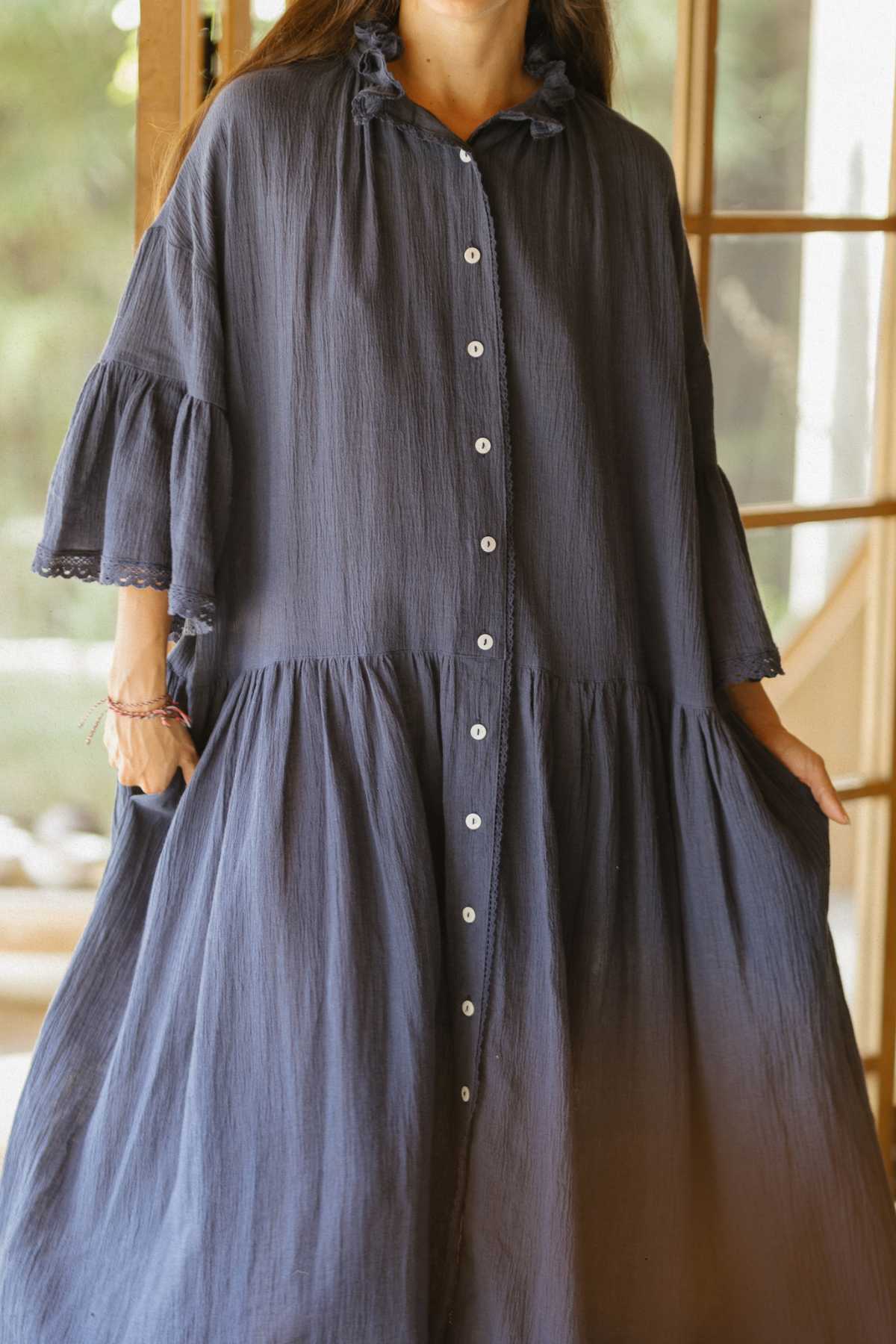 A person stands indoors near a window, elegantly dressed in The Juliette Linen Gown by Myrah Penaloza. This loose, flowy gown showcases a button-down front and features a ruffled, tiered design with lace detailing on its long sleeves. The softly blurred background enhances the graceful scene.