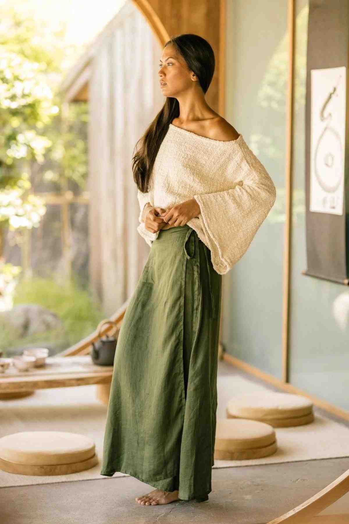 A person stands in a sunlit room, wearing the Spirit Top and Shanti Wrap Skirt Set from Myrah Penaloza. The off-shoulder white sweater and long, adjustable green skirt are made from 100% linen. They are barefoot, looking to the side with a contemplative expression. The background features wooden elements, round cushions, and minimalist decor with natural light.