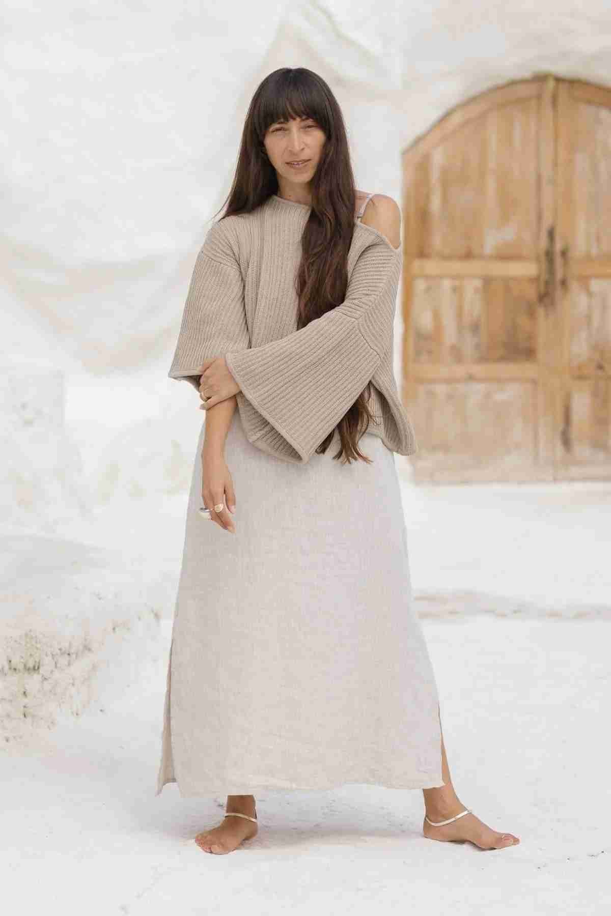 A woman with long, dark hair stands barefoot on a light, sandy surface. She is wearing the luxurious Spirit Top Sweater Knit from Myrah Penaloza, an elegant beige off-the-shoulder long-sleeved sweater made from cotton fabric, paired with a matching long skirt in light tones. The background showcases a rustic wooden door and white walls.